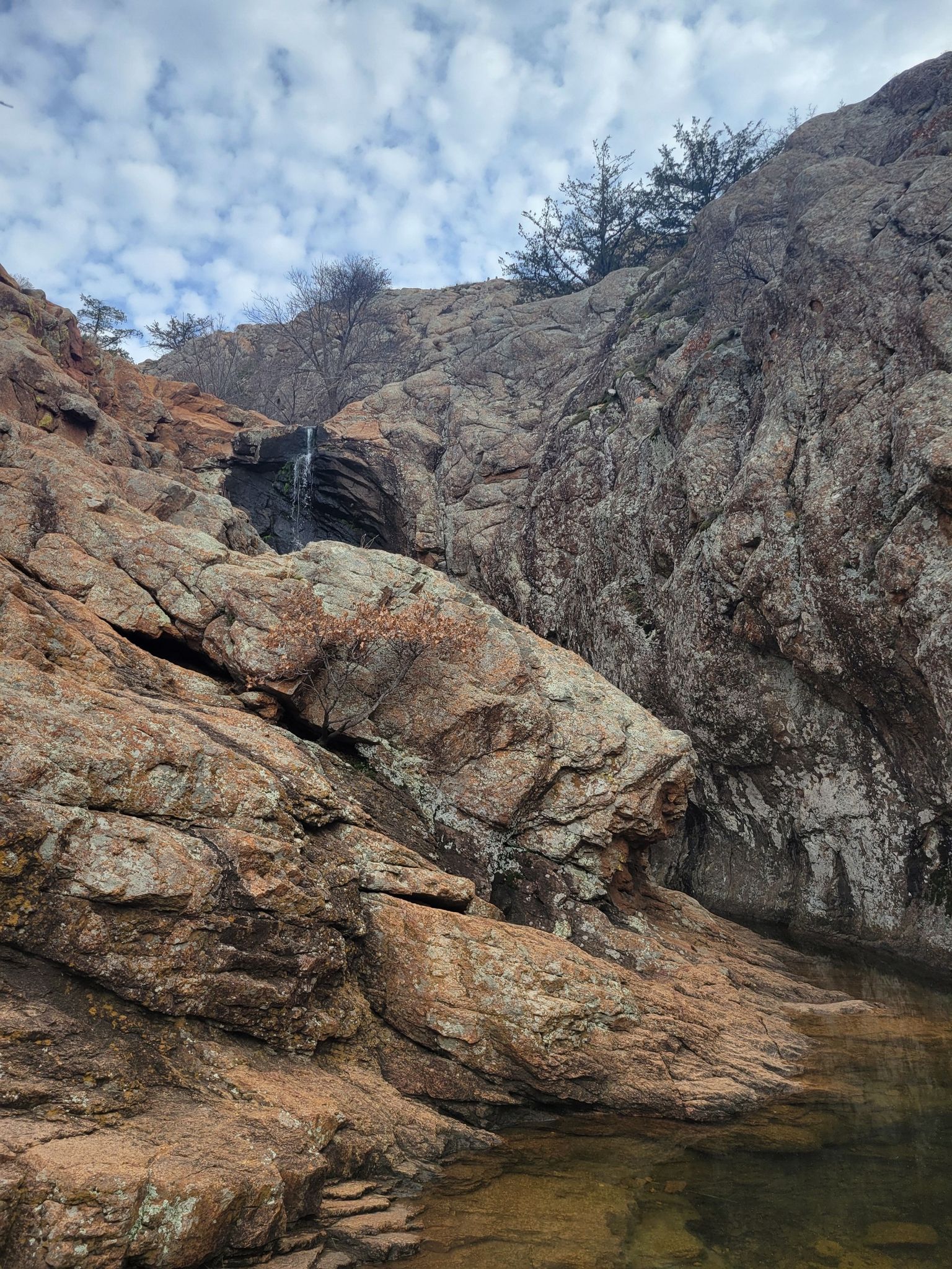 A small waterfall cascades down rugged, rocky cliffs into a dark pool. - Post Oak Falls Trail