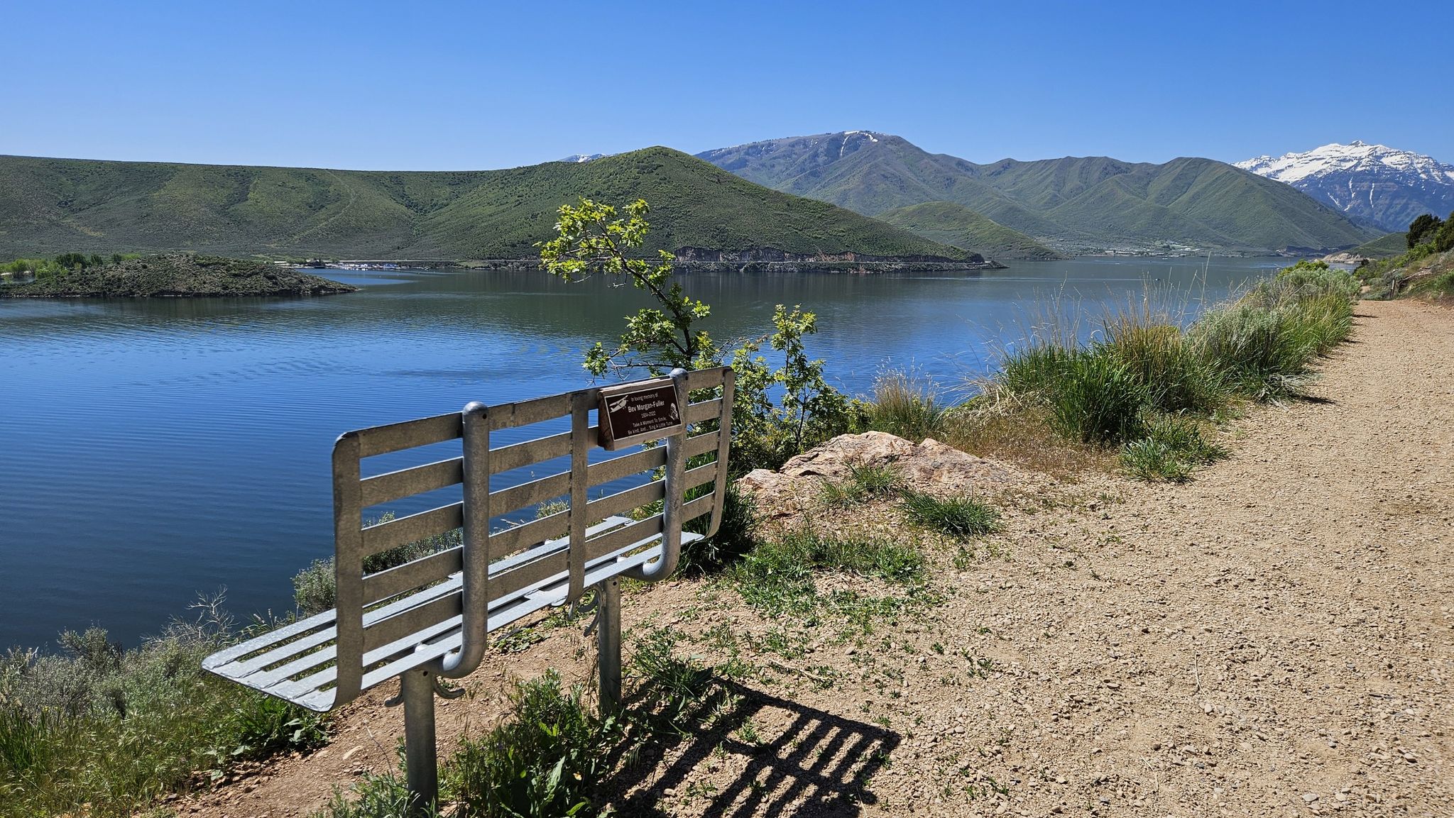 A metal bench by a dirt path overlooks a large lake and green mountains. - Deer Creek Trail