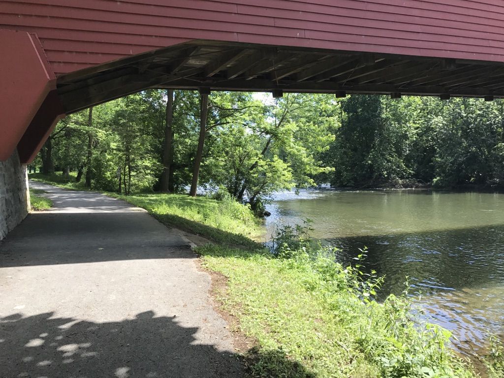 View from under a red bridge, showing a path, forest, and calm water. - Maiden Creek loop