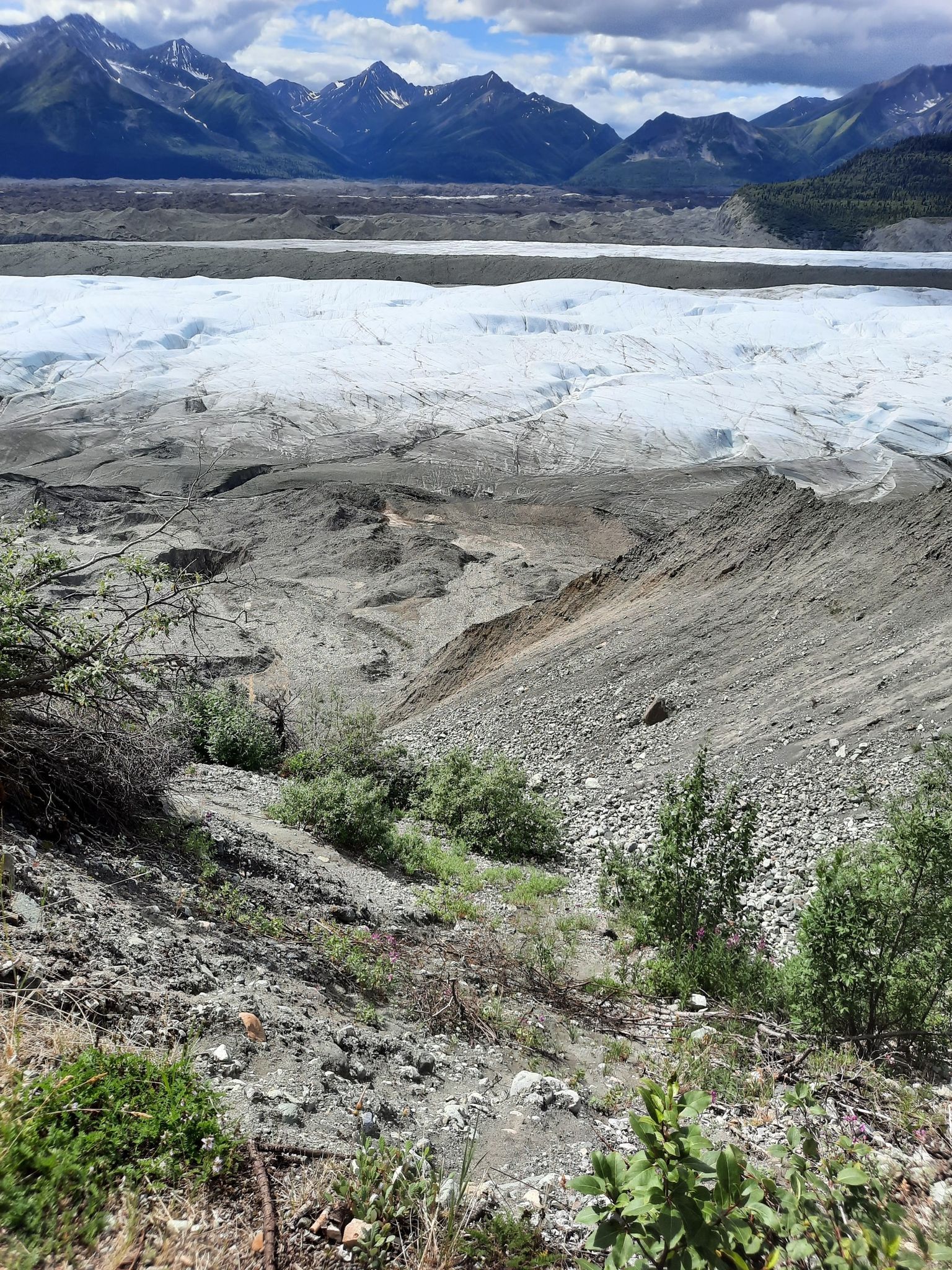 A vast glacier and rocky terrain under a cloudy sky with mountains. - Erie Mine Trail