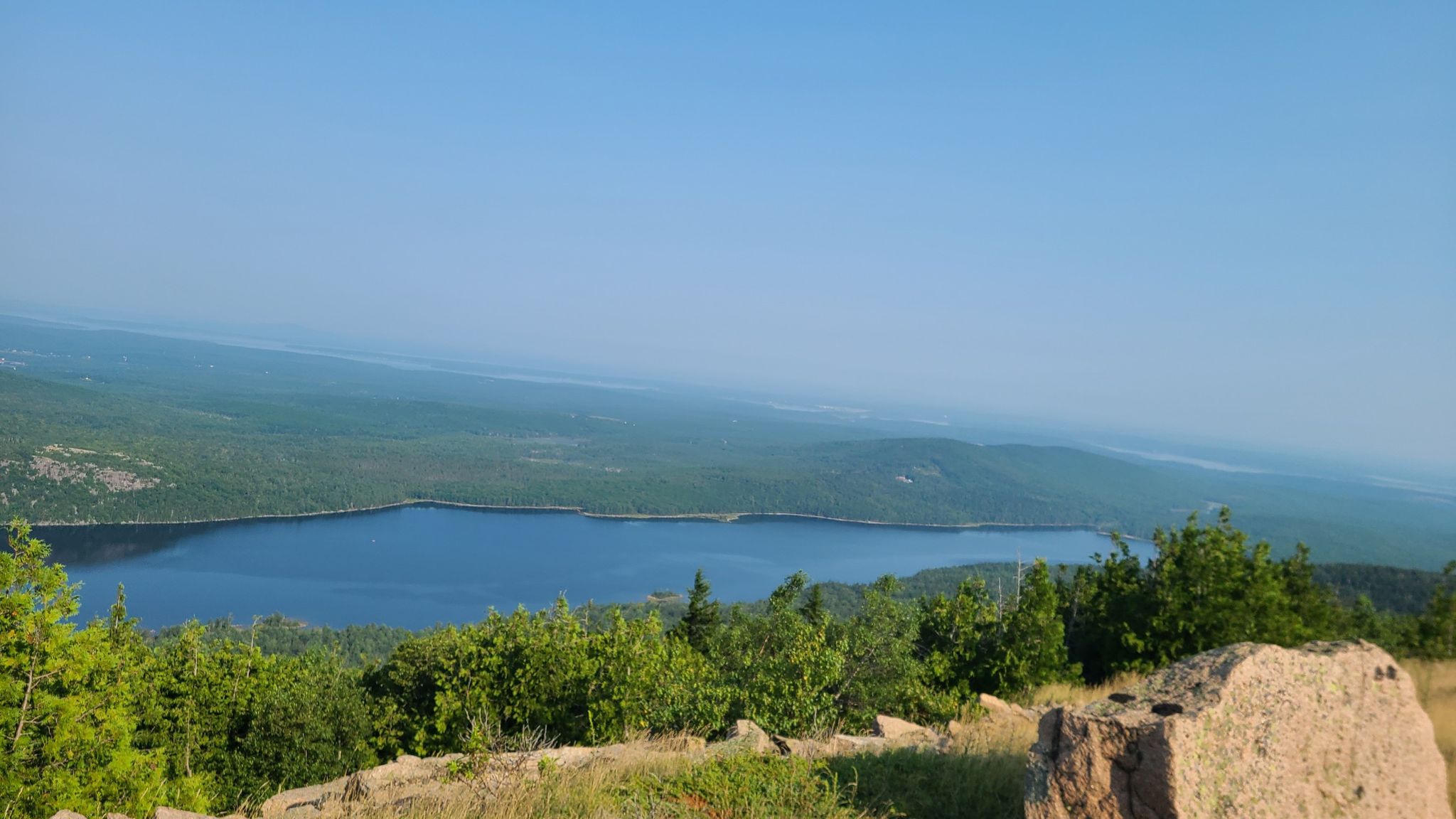 Panoramic view of a calm lake surrounded by green forests under a clear sky. - Cadillac Mountain from the Hulls Cove Visitor Center