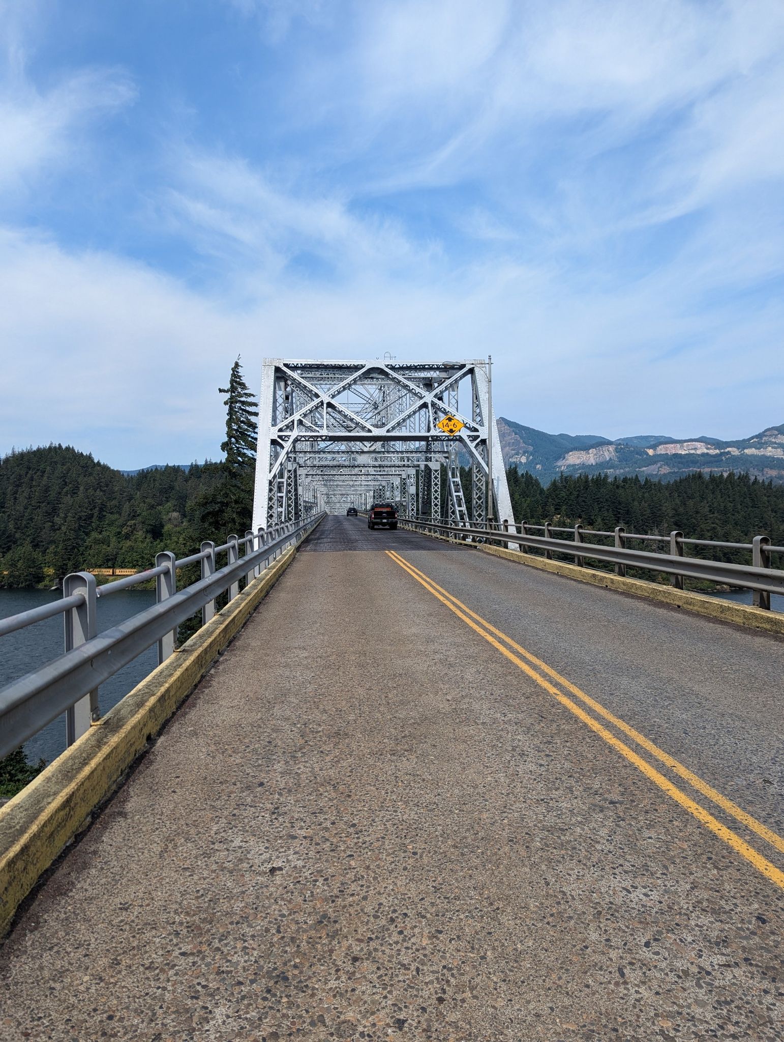 A large metal truss bridge spans a river, with cars and forested mountains. - Historic Columbia River Highway State Trail