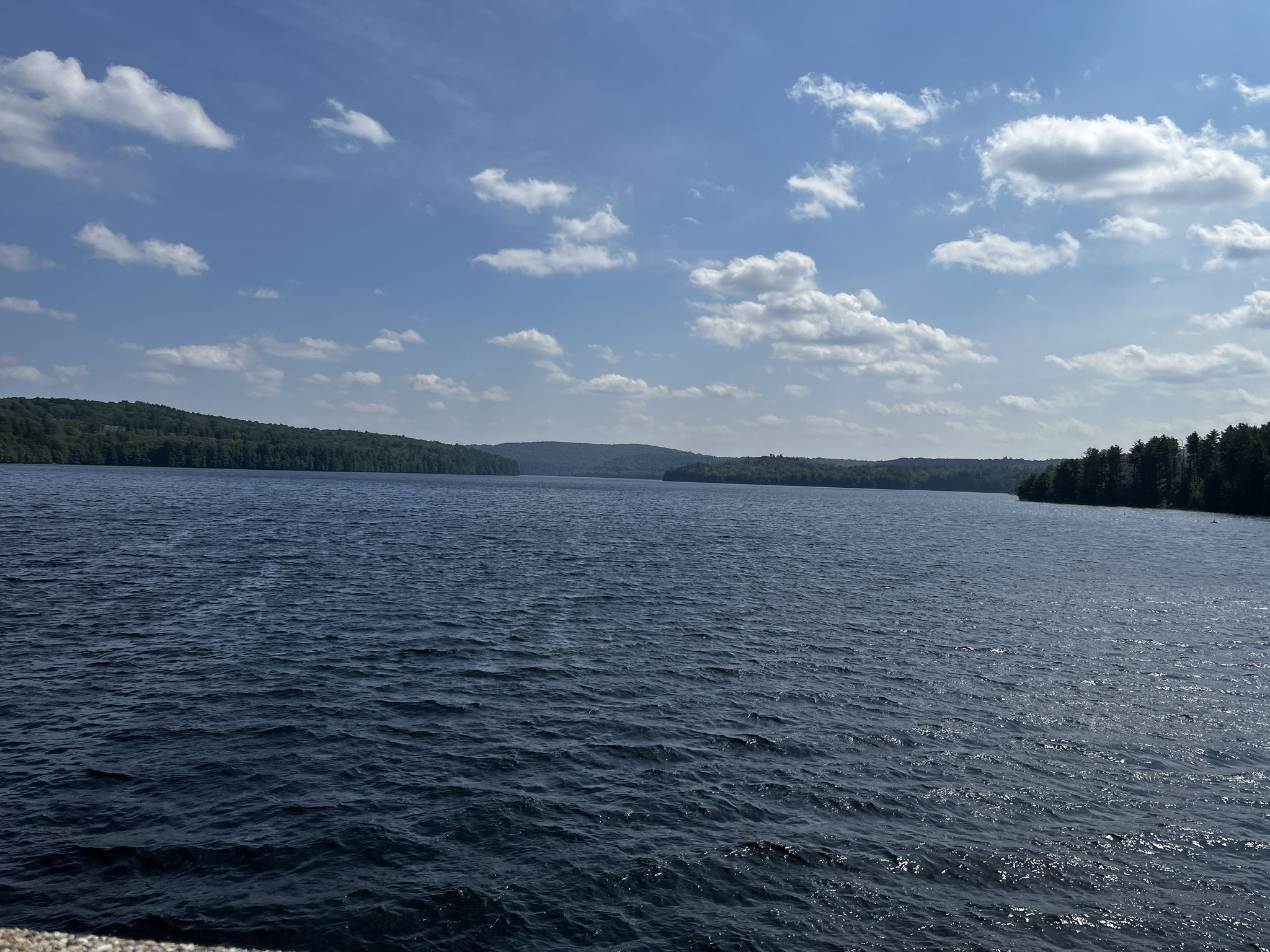 Wide shot of a large lake with forested shores under a cloudy blue sky. - Nepaug Reservoir Trail