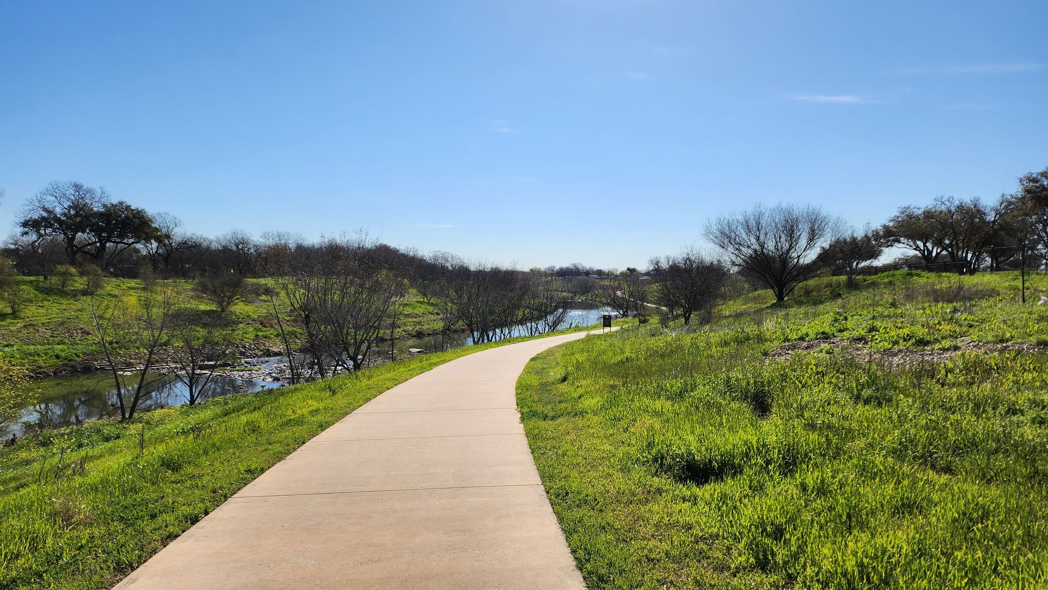 Paved path winding through green grassy hills beside a river, sunny day. - Mission San Juan Capristrano – Mission Concepción loop from San Antonio