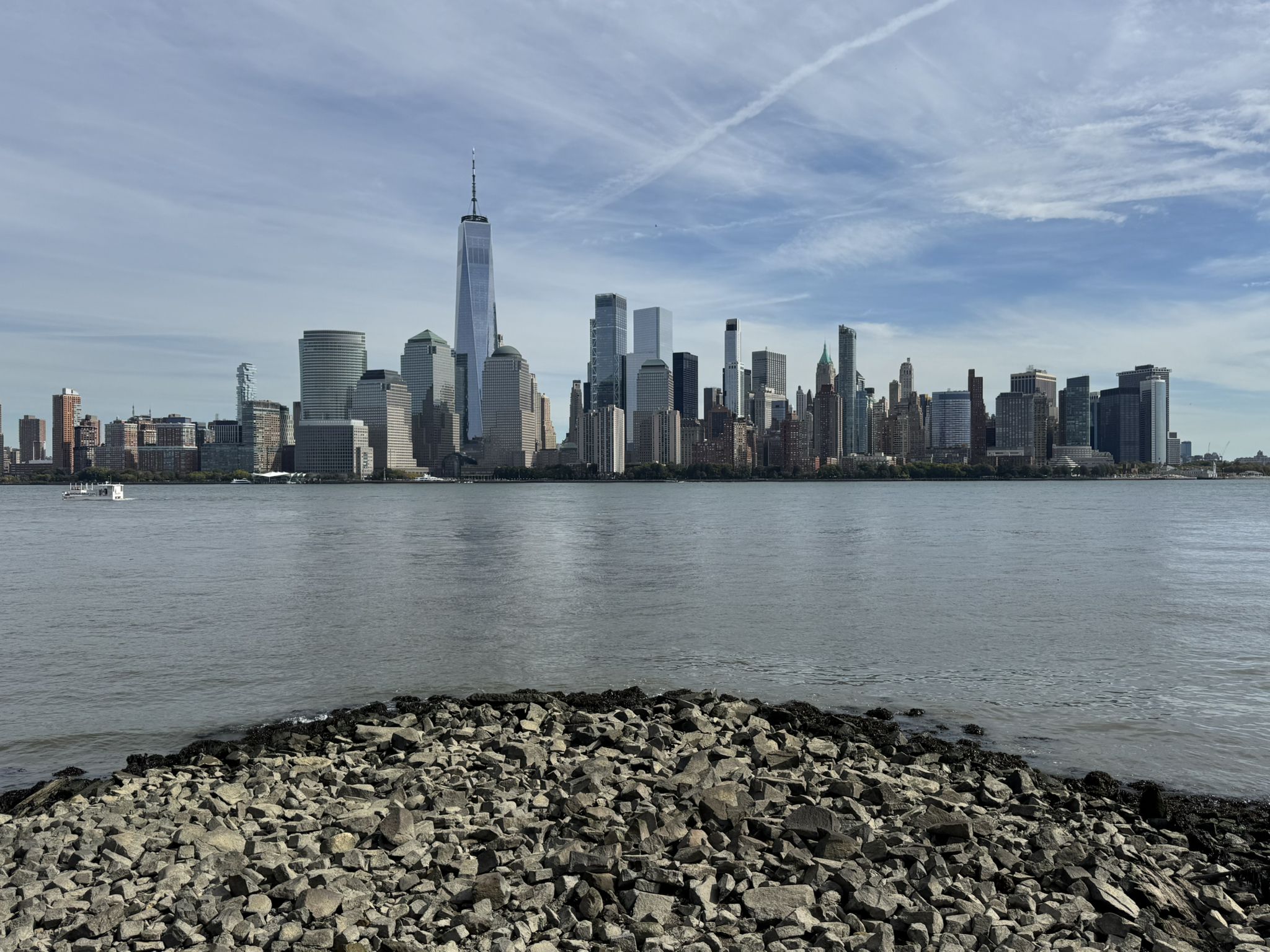 Distant city skyline across water with a rocky foreground under a cloudy sky. - Hudson River Waterfront Walkway