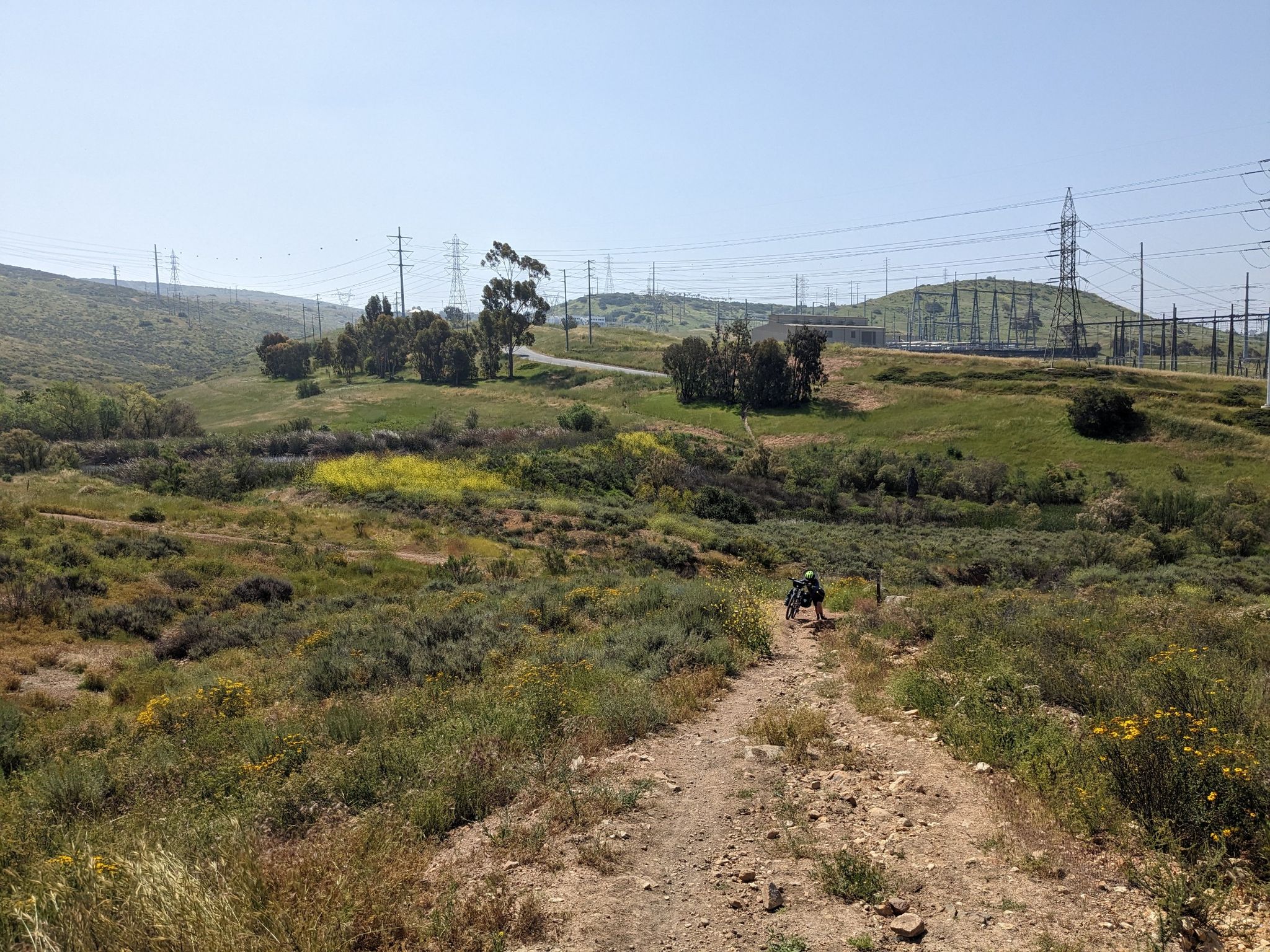 A dirt trail winds through green hills with power lines and a distant cyclist. - Rock House Trailhead – Tiki Hut loop from Mount San Miguel Park