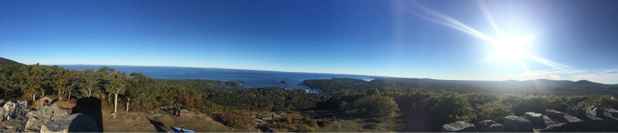 Wide panoramic view of a sunlit coastline, forest, and distant hills. - Mount Battie via Tablelands and Nature Trail