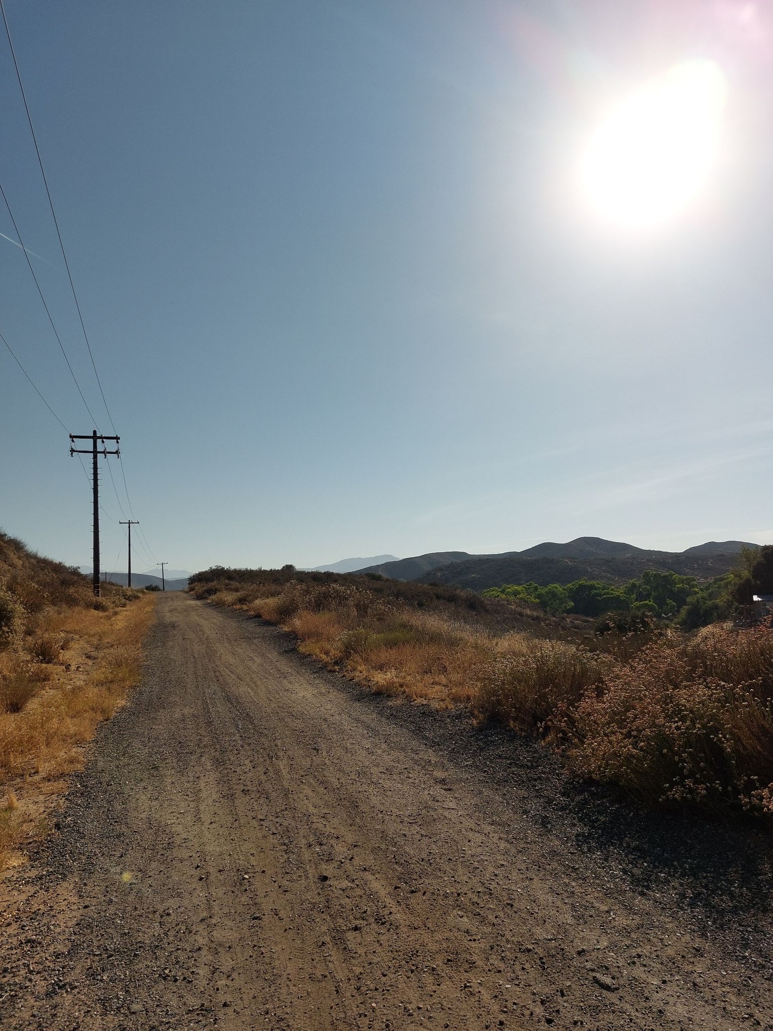 Dirt road with power lines, leading towards a bright sun and hills. - Oak Mountain loop from Dripping Springs