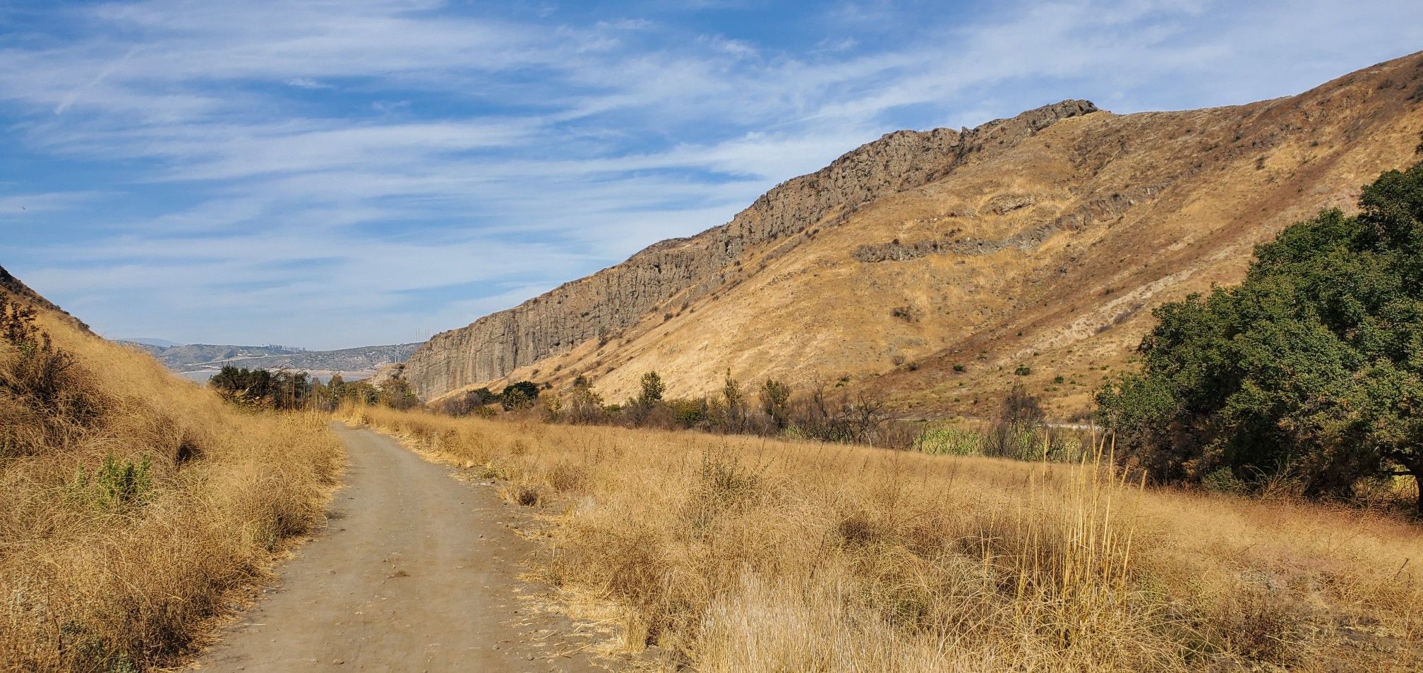Dirt road through dry grass, flanked by a steep rocky hill under a blue sky. - Canejo Canyon Open Space Loop