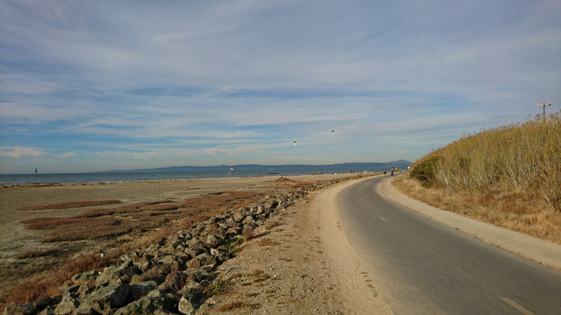 Paved path curving alongside a rocky shore and calm water under a partly cloudy sky. - San Francisco Bay Trail: Burlingame to Redwood Shores