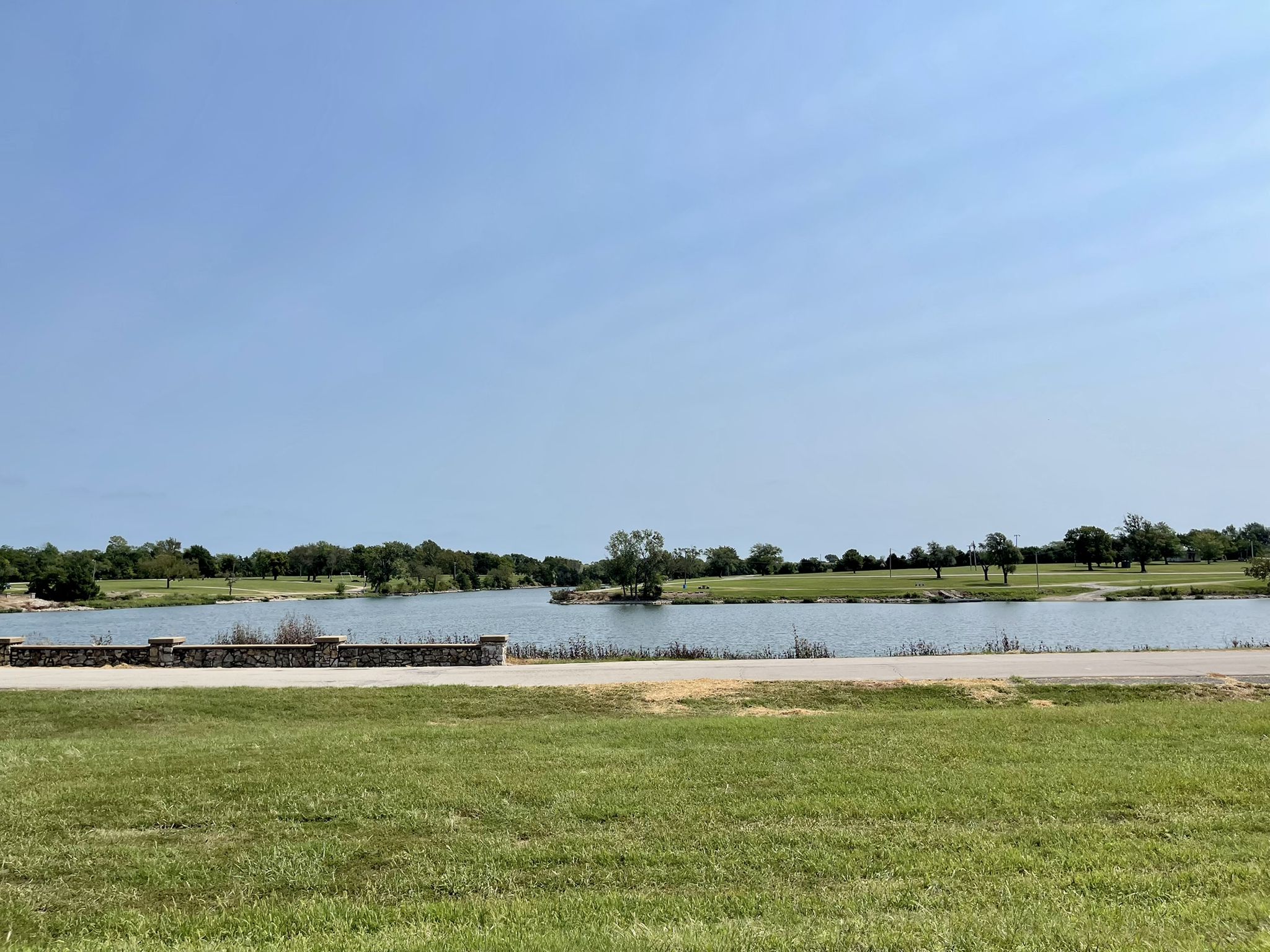 A wide lake surrounded by green grass and trees under a bright blue sky. - Prairie Spirit Trail