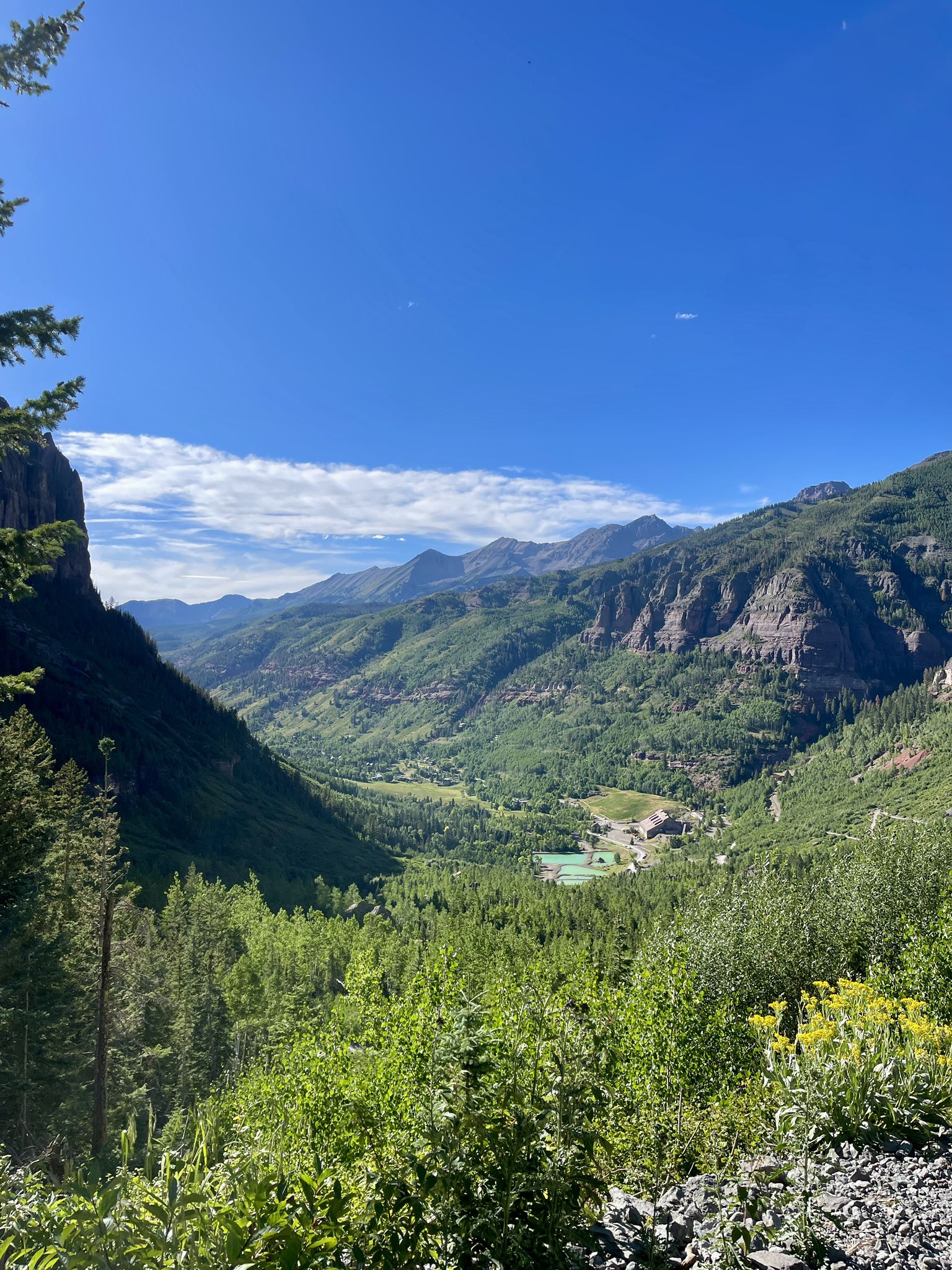 Expansive mountain valley view with green forests and distant buildings under blue sky. - Bridal Veil Falls, Black Bear Pass Loop