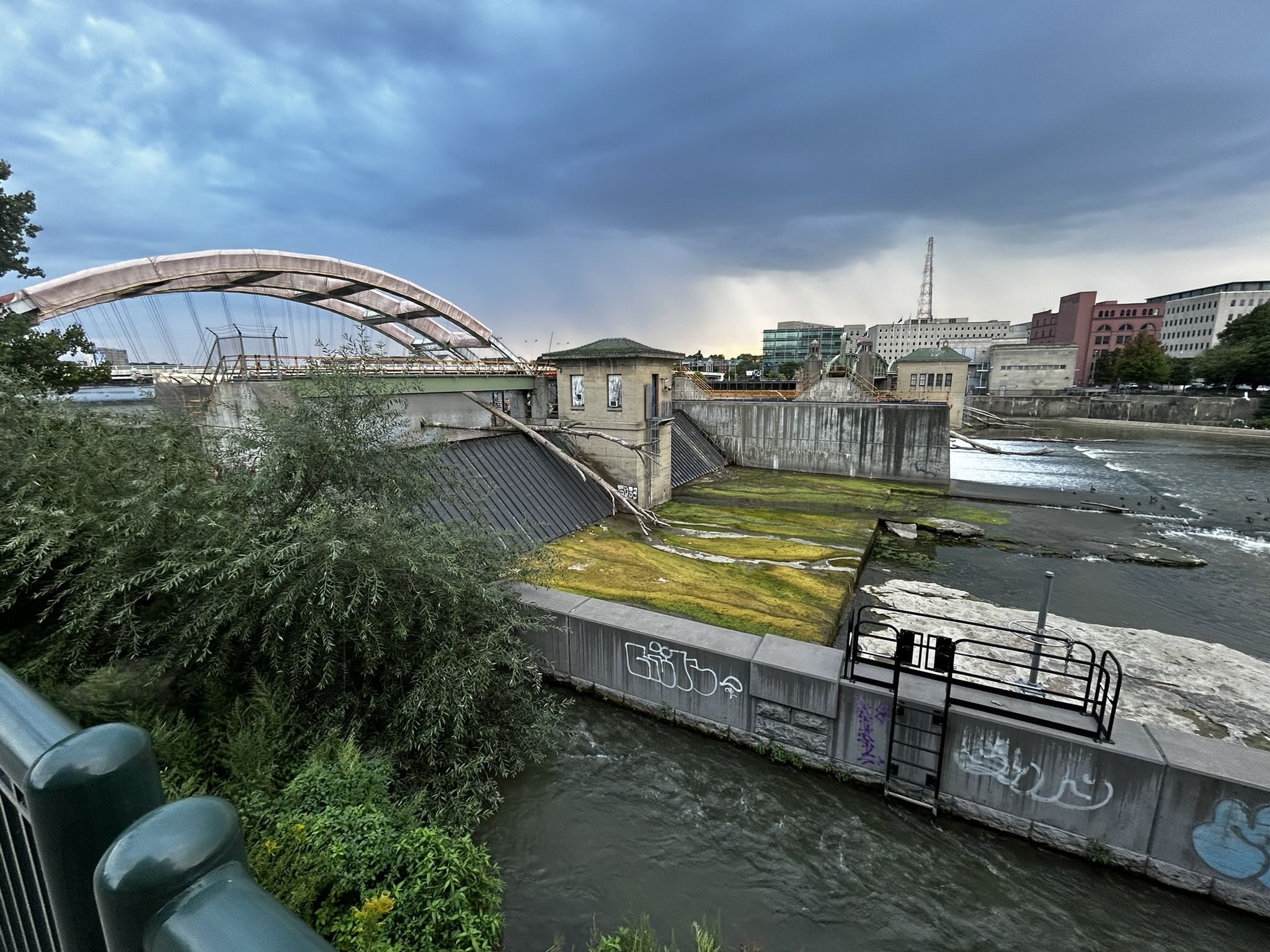 Overcast view of a dam, arched bridge, and urban river under dark clouds. - View of Court Street Dam loop from Rochester