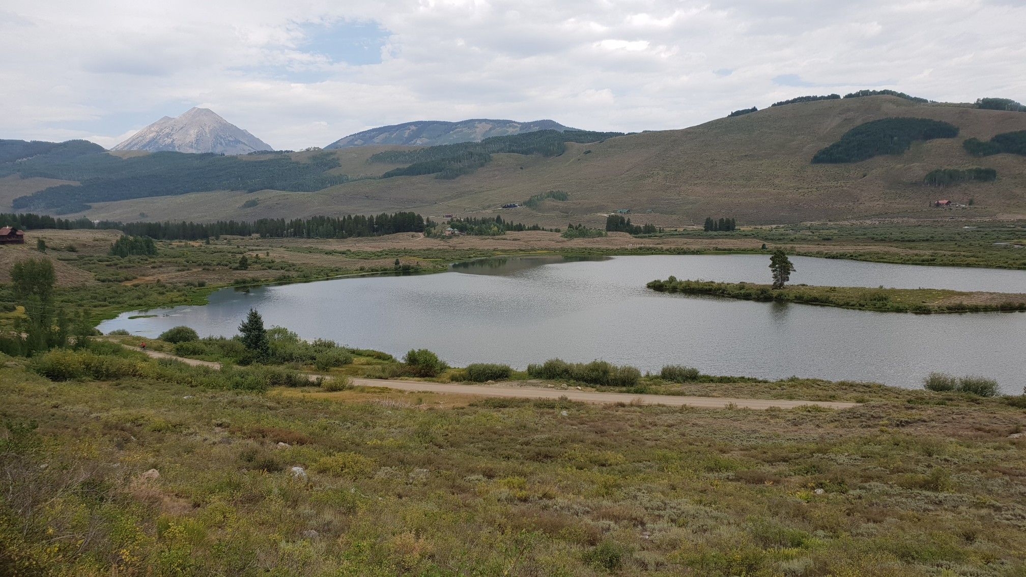 Calm lake surrounded by grassy hills and distant mountains under cloudy sky. - Peanut Lake – Crested Butte Downtown loop from Crested Butte
