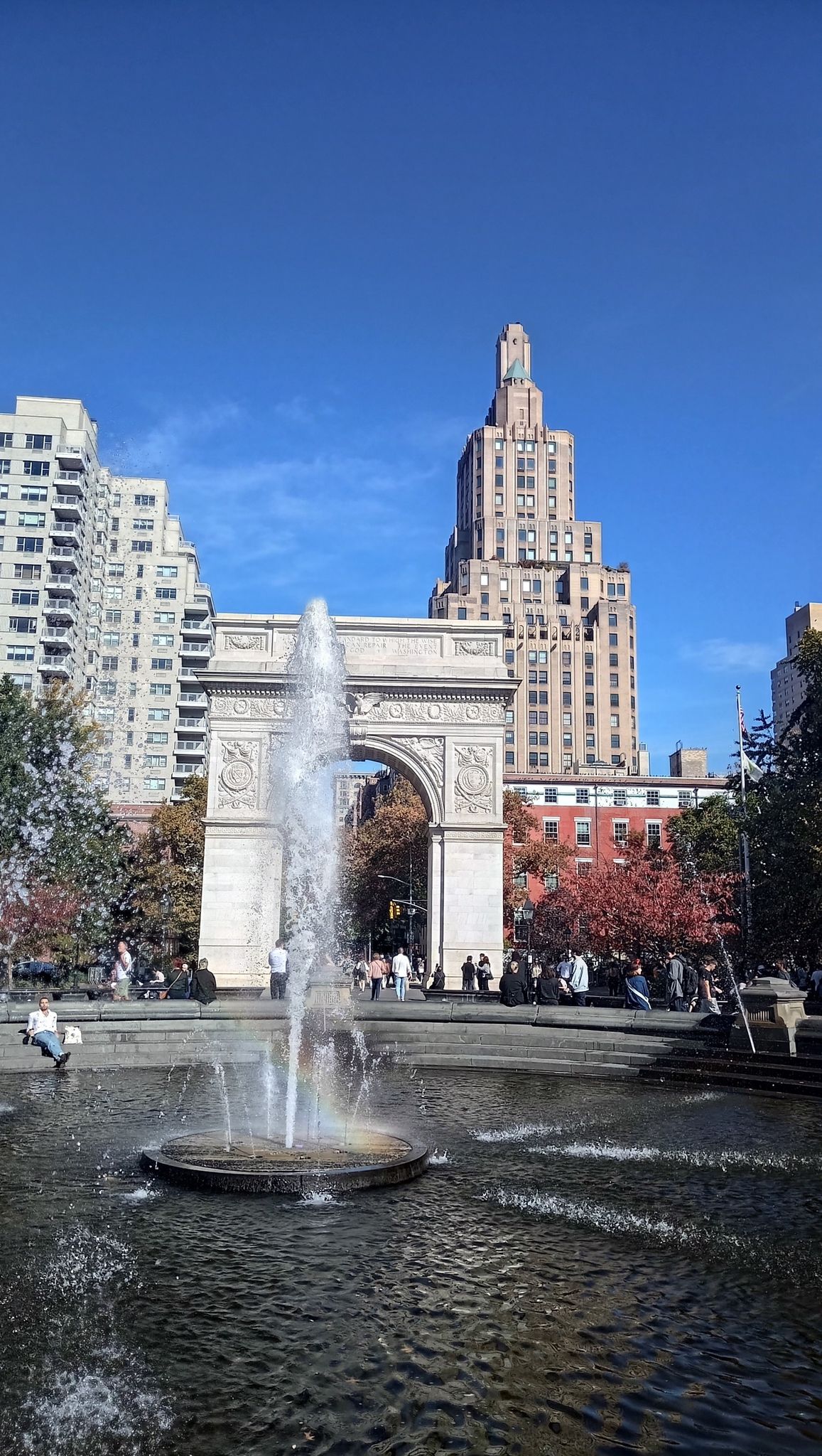 Bright daylight view of a fountain and archway with tall buildings and autumn trees. - Washington Square Park Loop