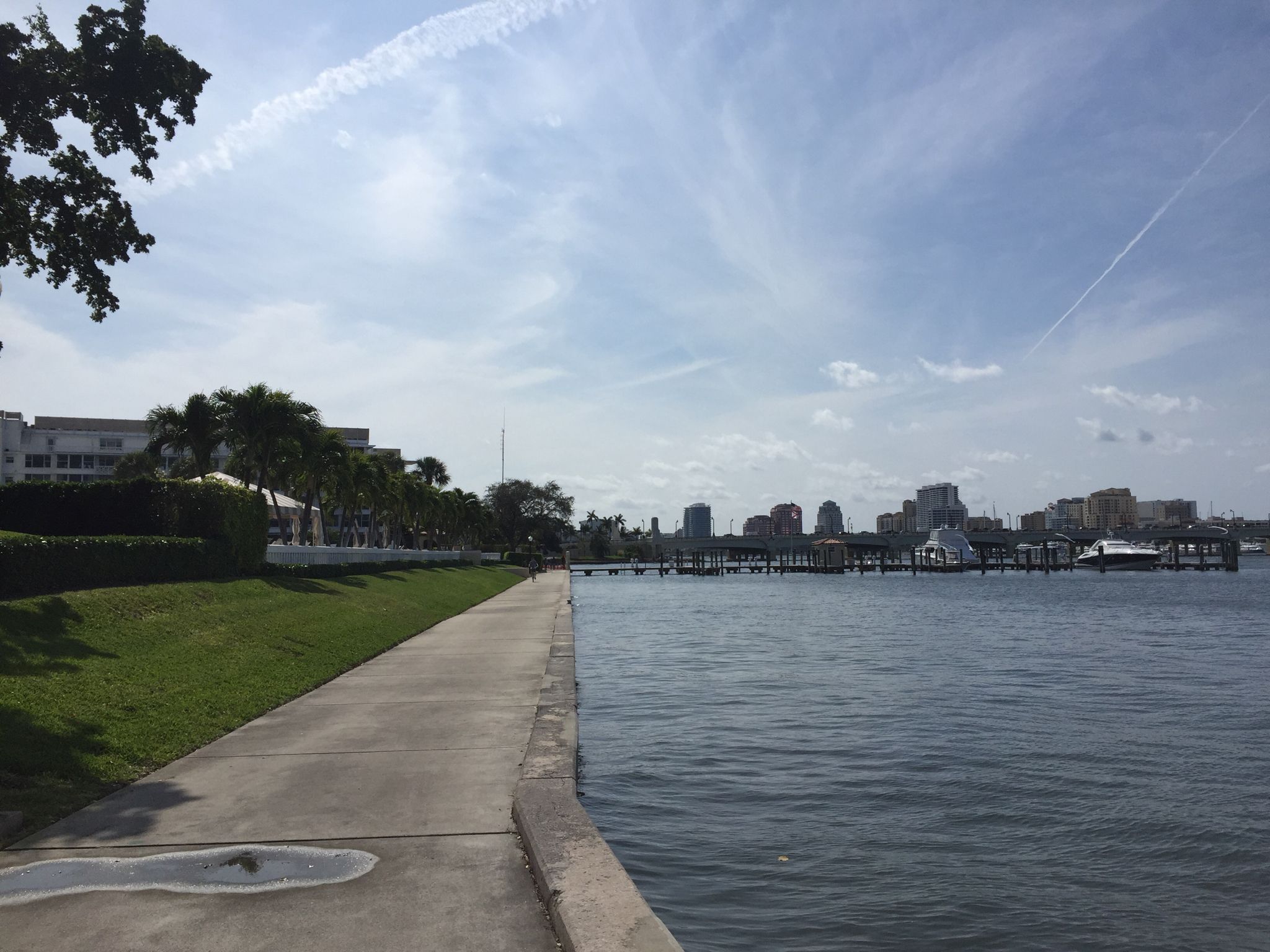 Concrete walkway beside calm water, with a distant city under a cloudy sky. - Running loop from West Palm Beach