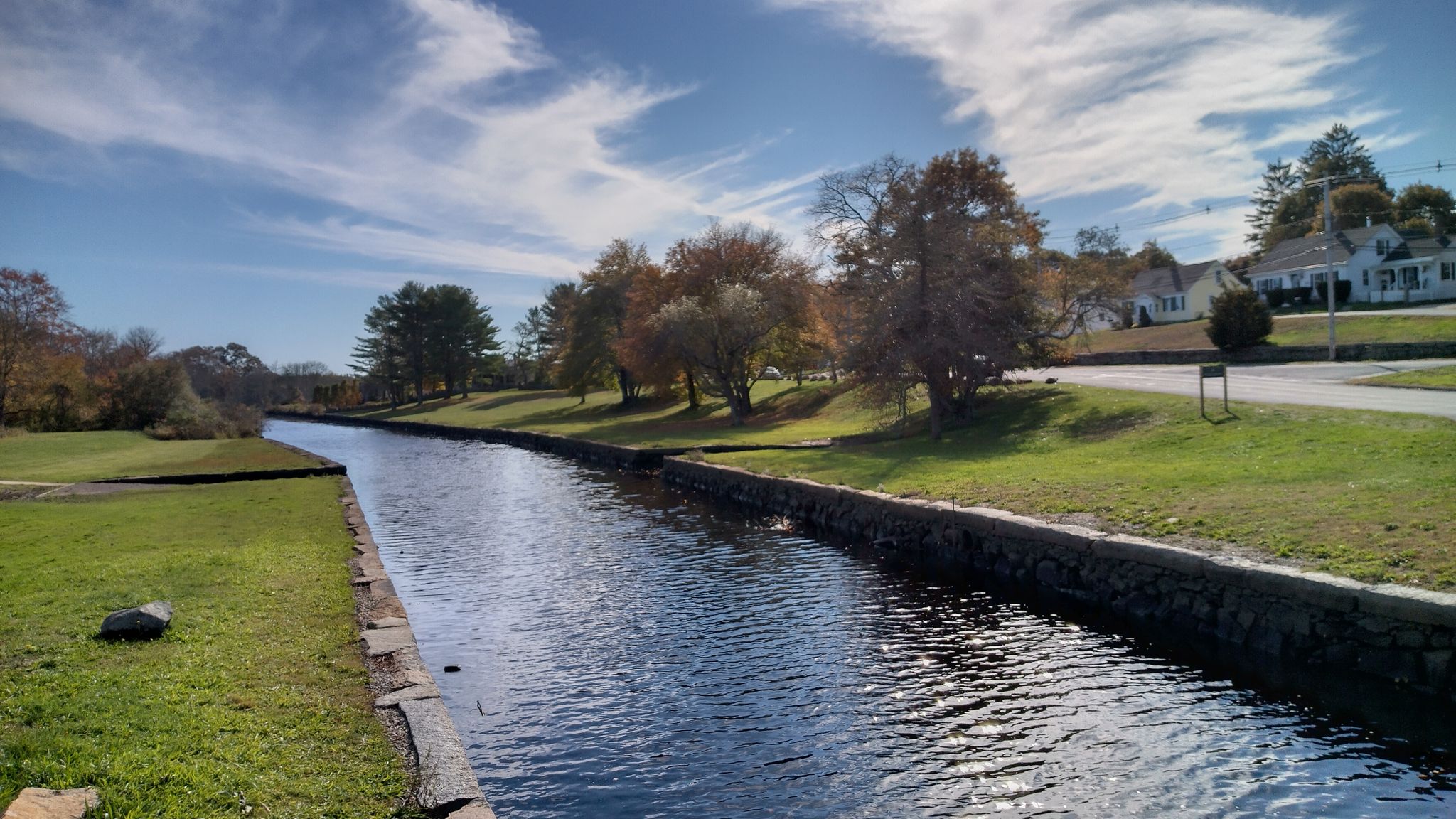 A narrow canal with stone walls, green grass, and houses under a blue sky. - Seapowet Marsh Management Area – Acoaxat Beach loop from Dartmouth
