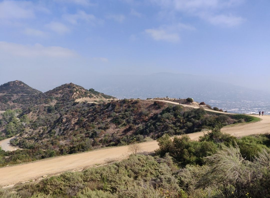 Winding dirt road through brush-covered hills with distant hazy city views. - Griffith Park loop