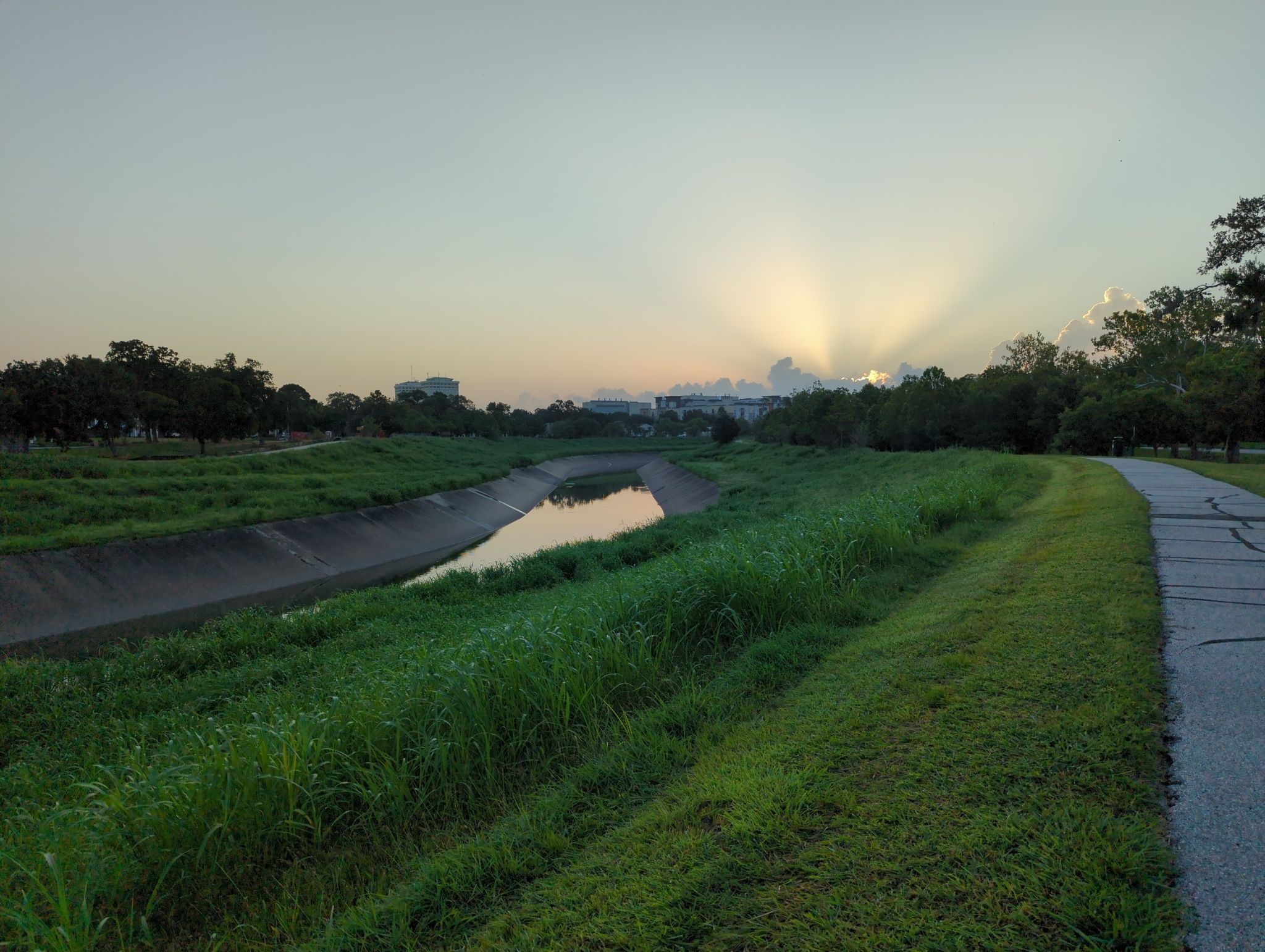 Sun rays pierce through clouds over a canal, green banks, and paved path. - Brays Bayou Greenway