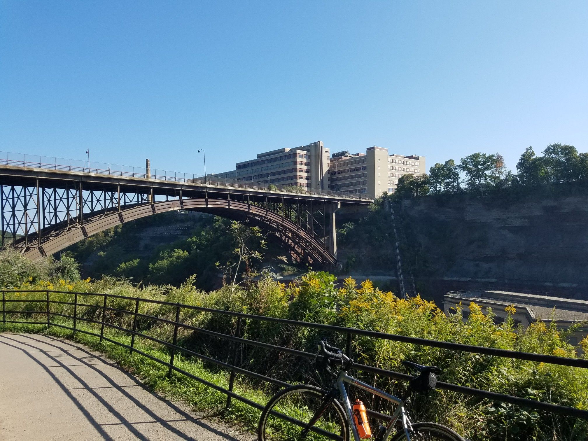 A bicycle on a path overlooks an arched bridge and buildings under a clear sky. - Lower Falls Overlook Trail