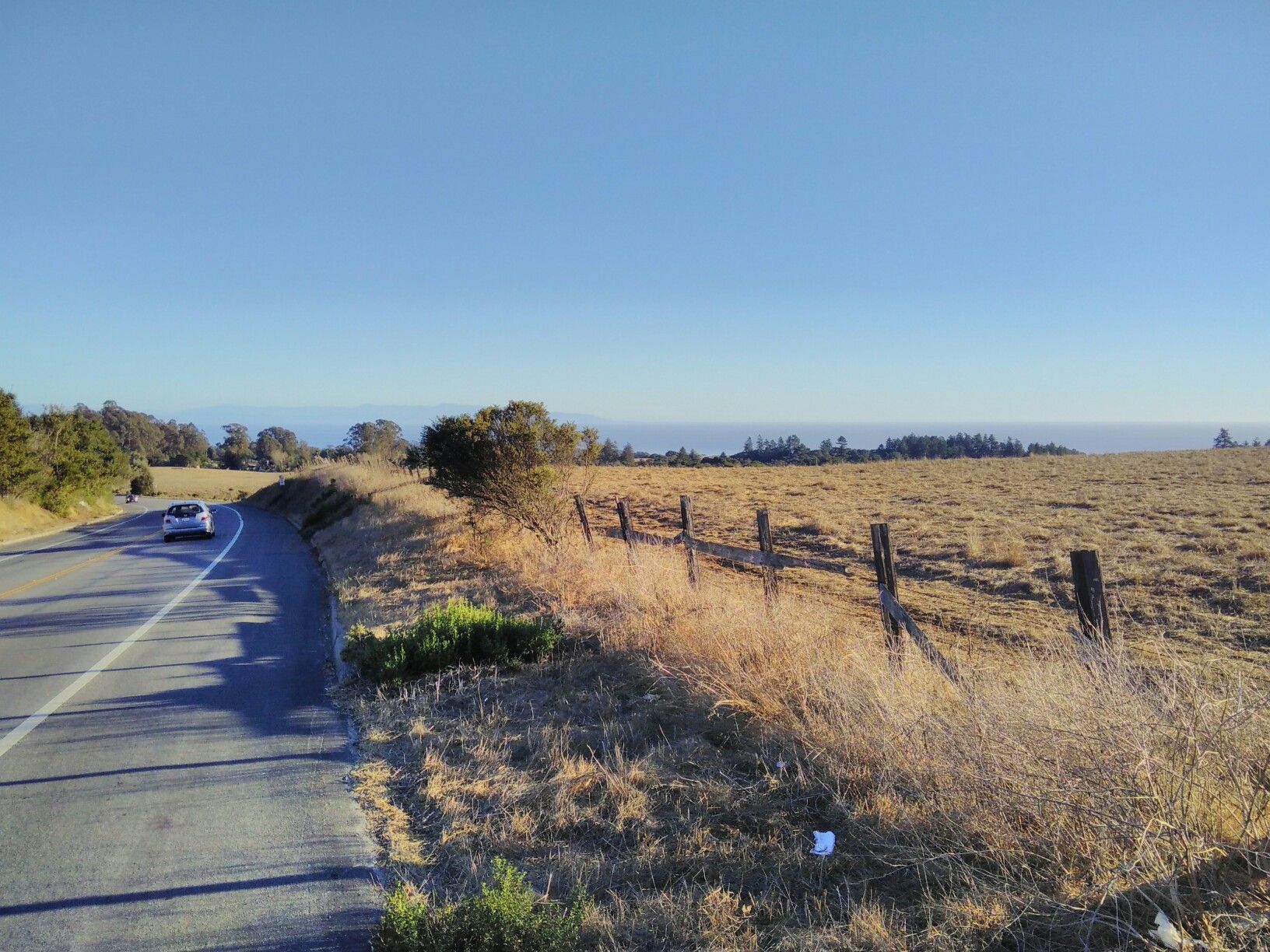 A car drives on a road beside a dry field with a fence, under clear sky. - Boulder Creek loop