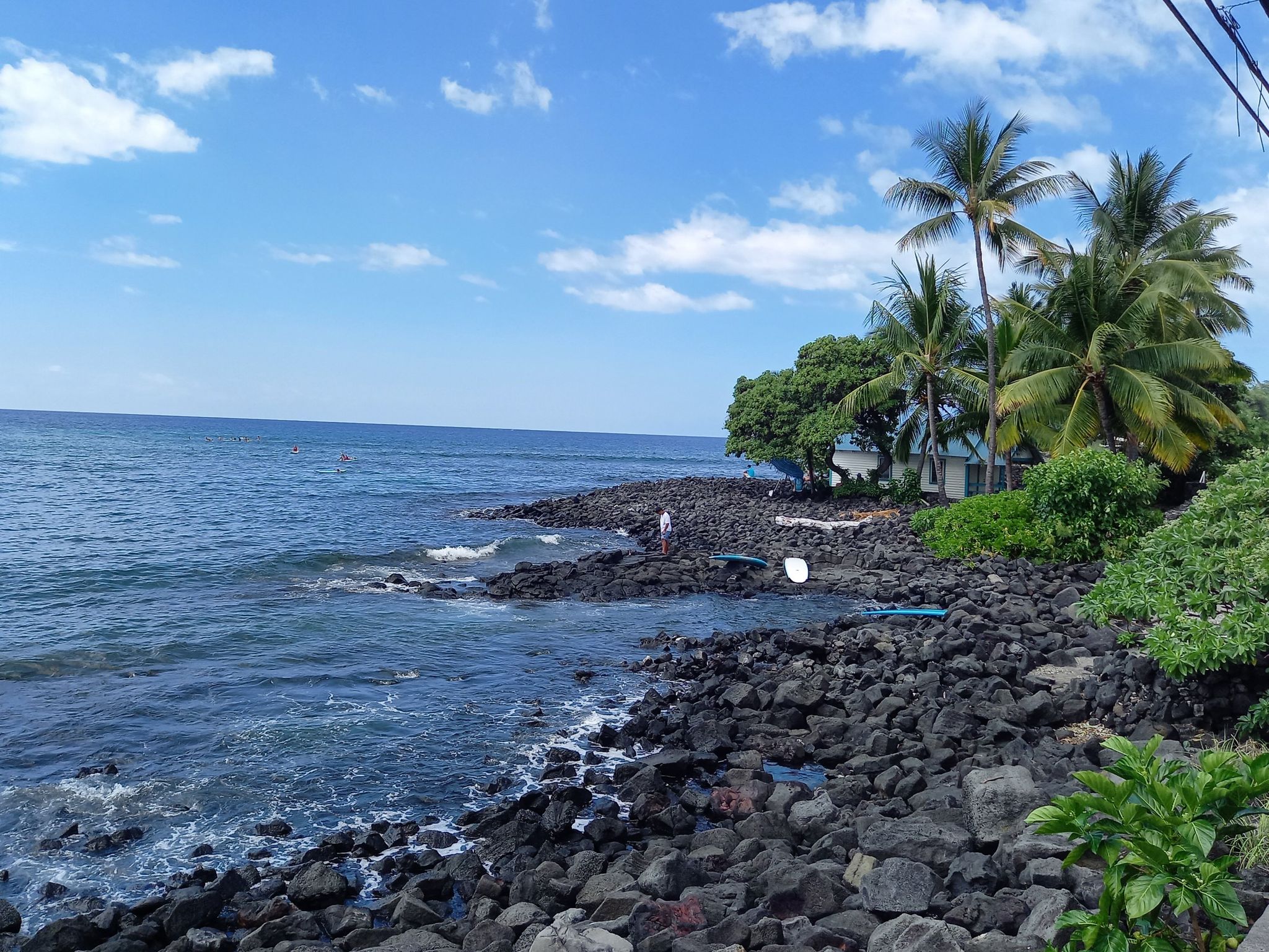 Sunny rocky coastline with blue ocean, palm trees, and a distant person. - Kahaluu Beach Park loop from White Sands Beach Park