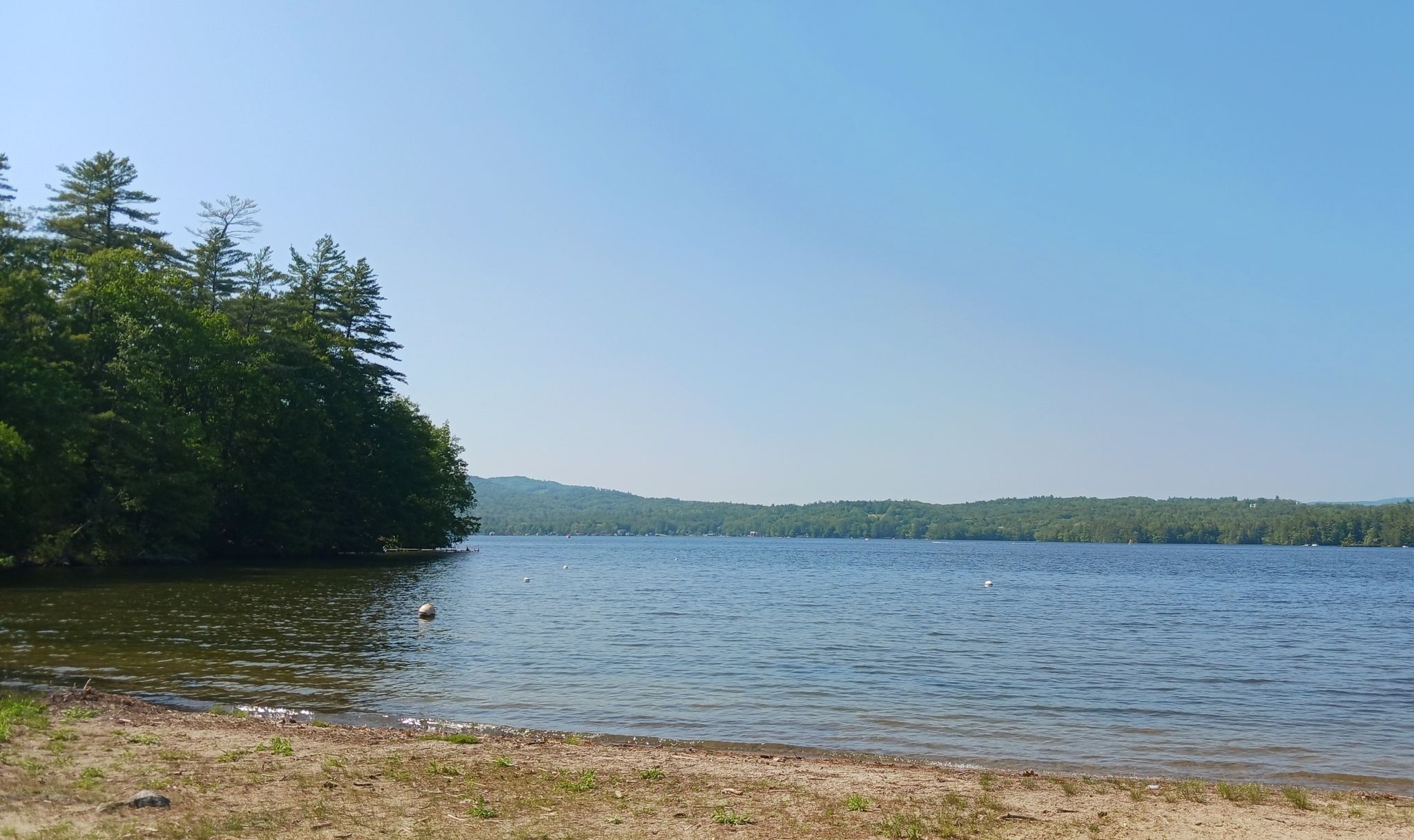 A wide lake with a sandy shore, forested hills, and a clear sky. - Webster Lake loop road run