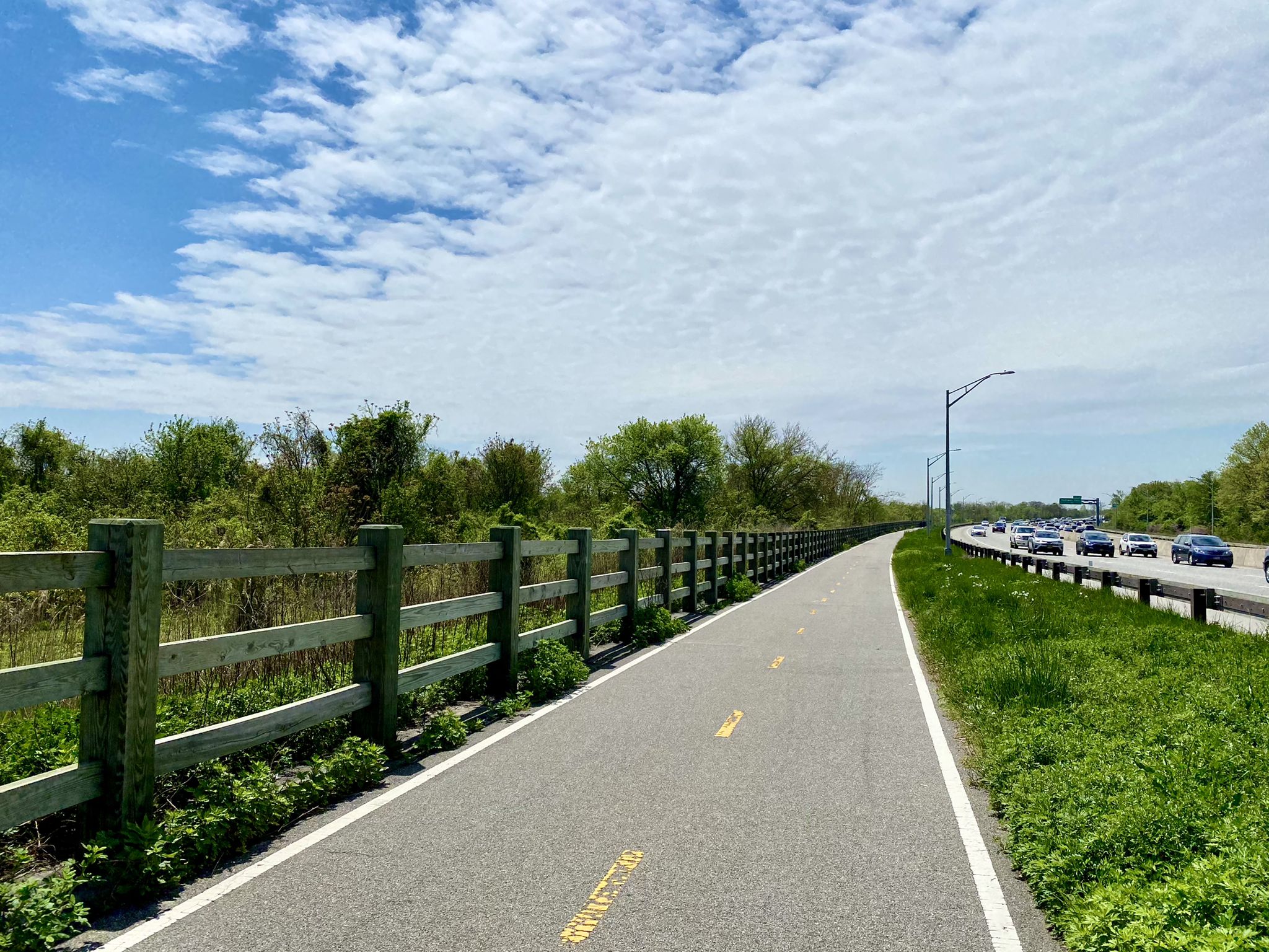 Paved path with wooden fence alongside a highway with cars. - Jamaica Bay Greenway – Jamaica Bay Greenway loop from Barren Island