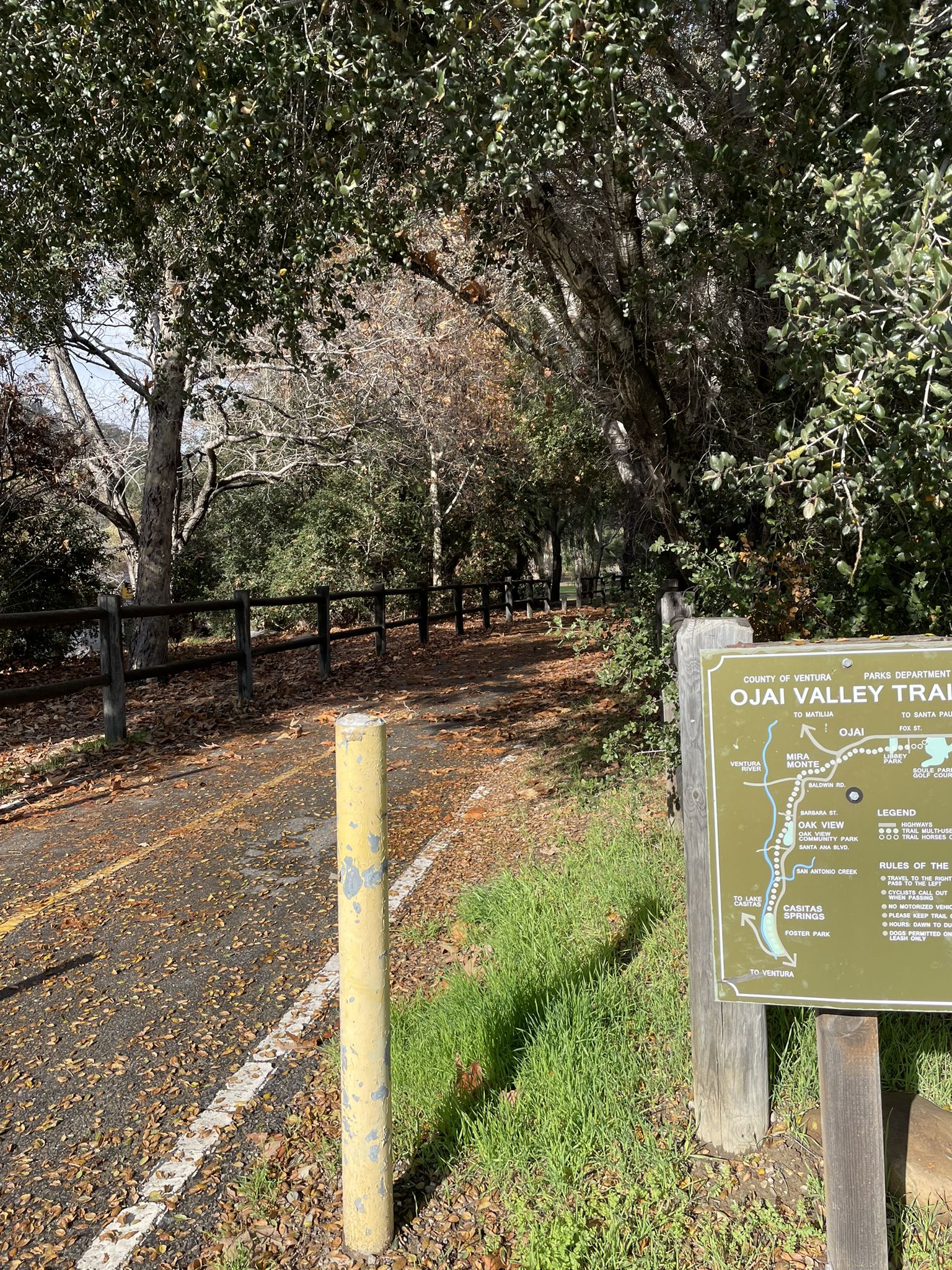 Paved path through dense trees, a wooden fence, and an information sign. - Ojai - Ventura Bike Path