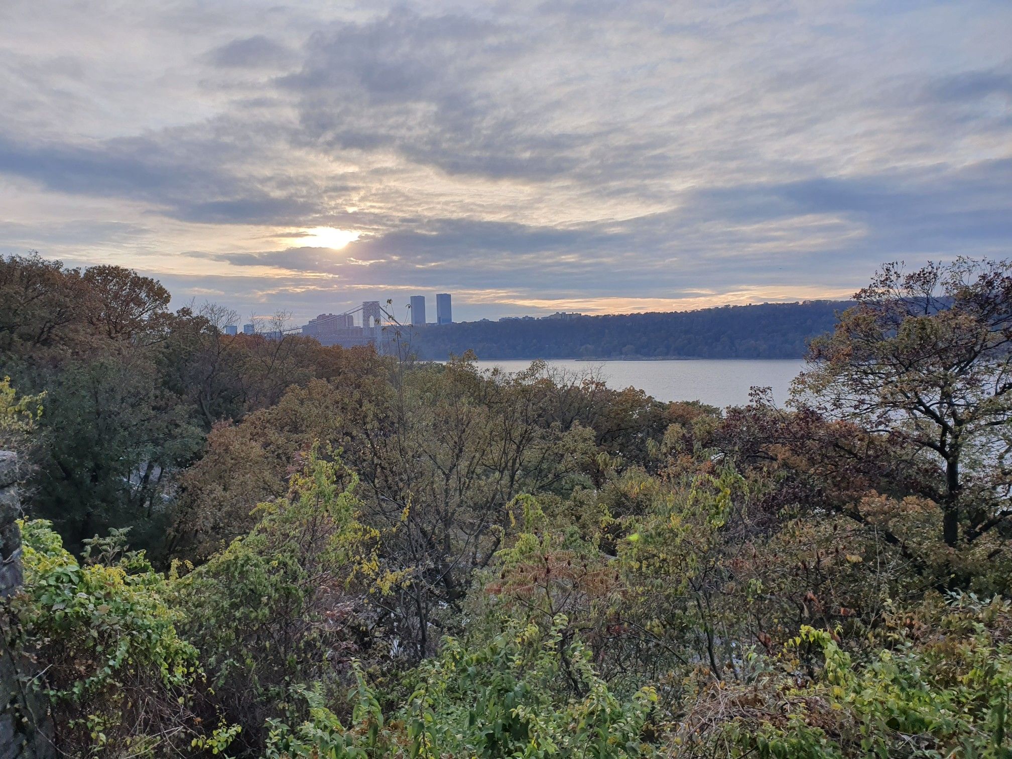 Sun peeking through clouds over a wide river, distant structures, autumn trees. - Fort Tryon Park Loop
