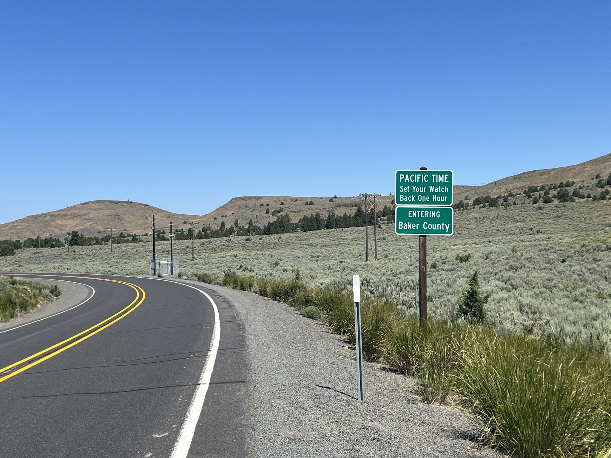 Paved road curves left past a green sign in a dry, hilly landscape. - Eldorado Pass via US26