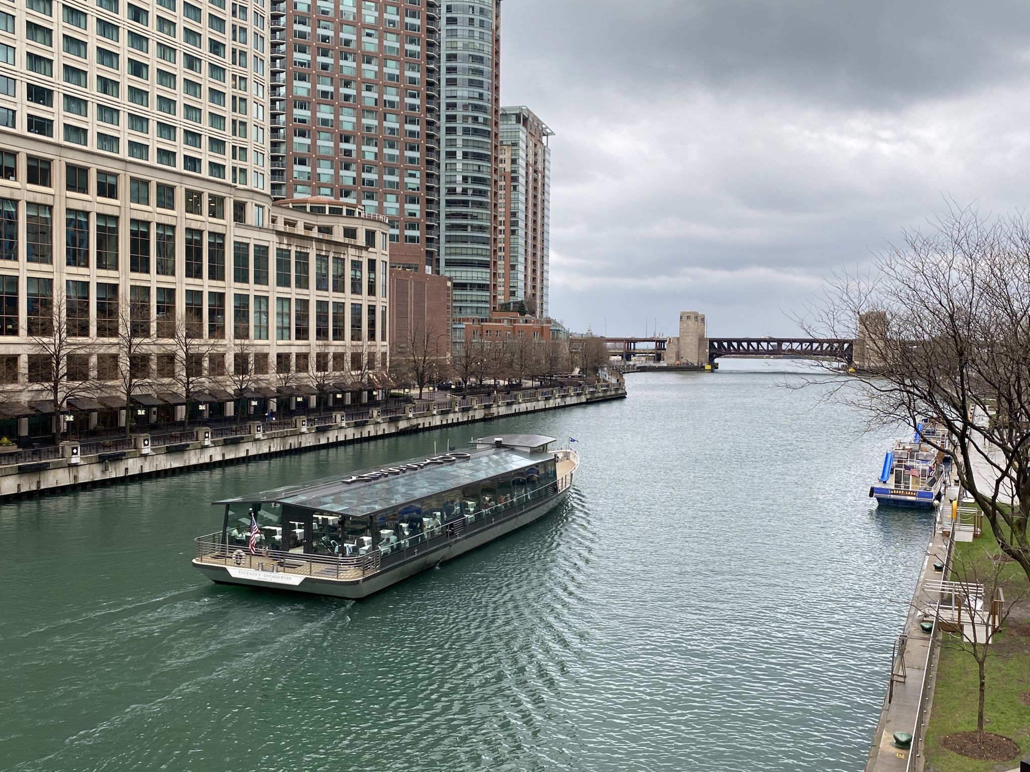 A tour boat navigates a river lined with tall buildings under an overcast sky. - Chicago Riverwalk