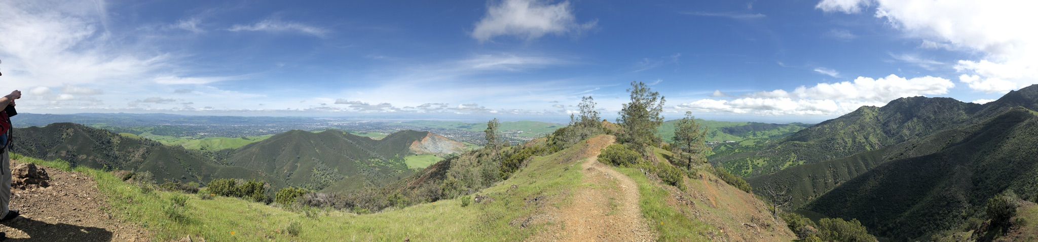 Panoramic view of a green valley and mountains under a partly cloudy sky. A person stands left. - Mitchell Canyon and Eagle Peak Loop