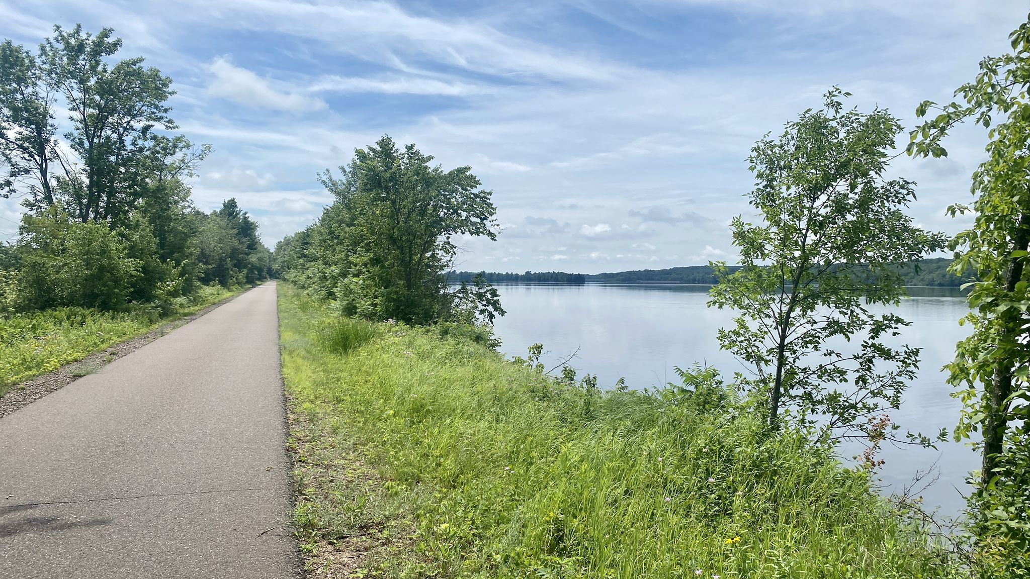 Paved path next to a wide lake, green trees, and a bright sky. - State Park Beach loop from Lake Hallie