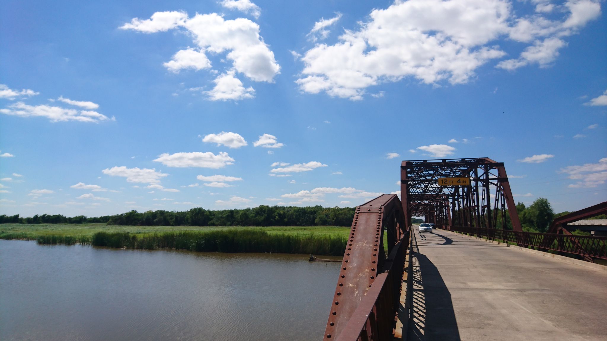 A rusty metal truss bridge crosses a wide river under a bright blue sky. - Lake Overholser Bridge – Lake Overholser Dam loop from First Indian Pentecostal Church