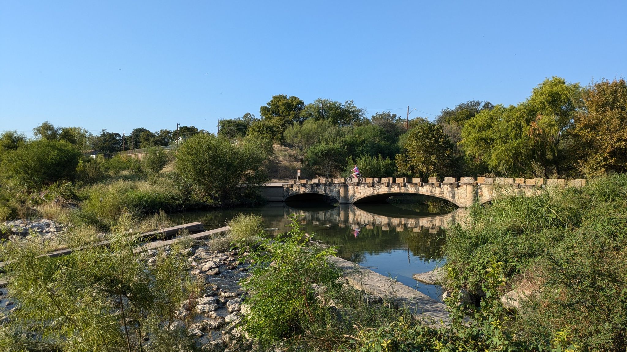 Sunny view of a stone arch bridge over a river with green foliage. - River Walk – Bexar County Courthouse loop from San Antonio