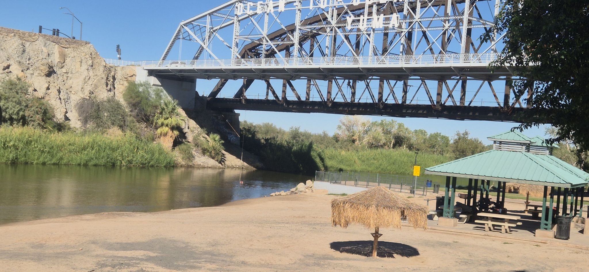 Sandy river beach with a thatched umbrella and picnic gazebo under a bridge. - East Wetlands & South Redondo loop