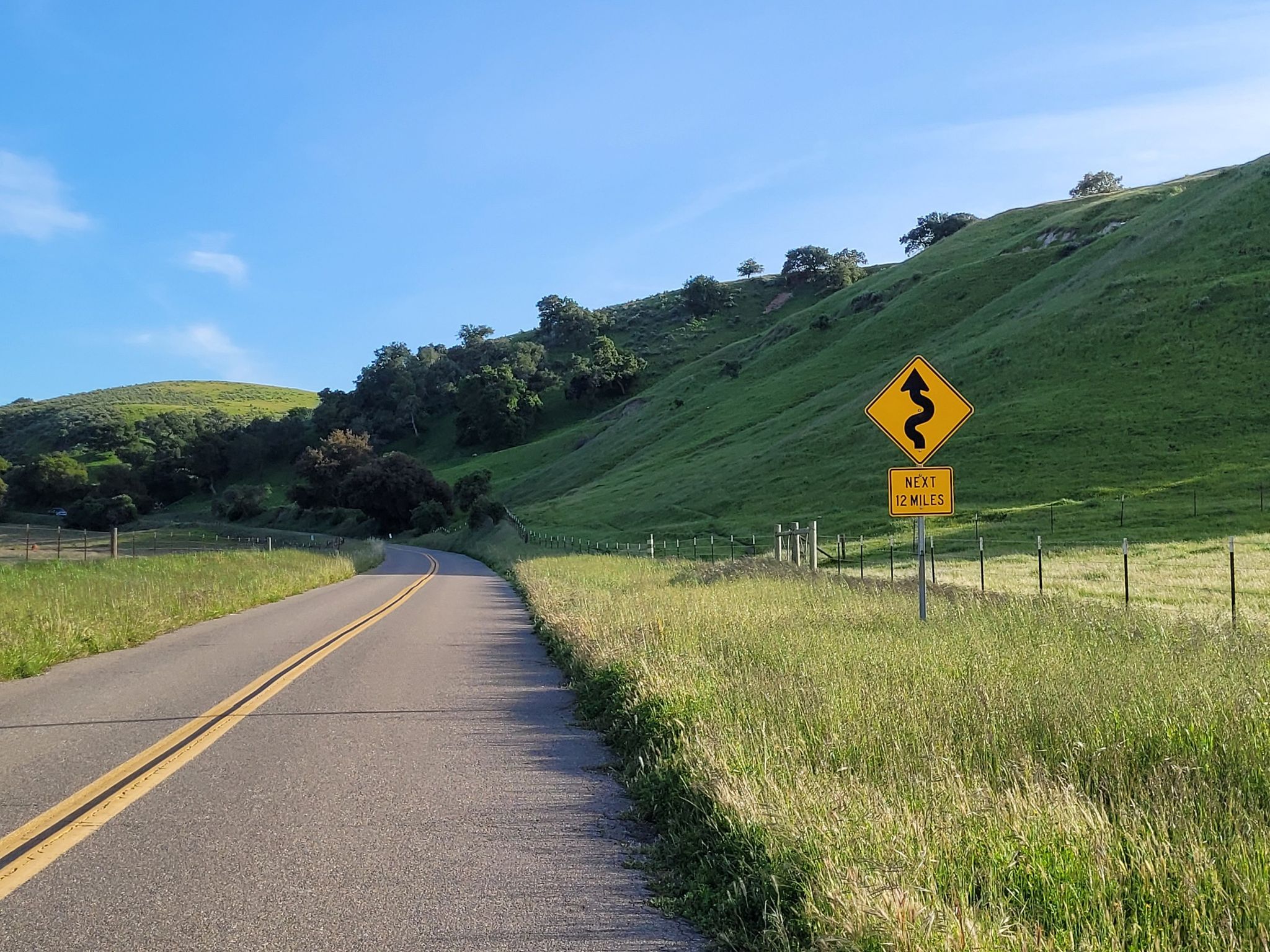 A paved road winds through green hills under a bright blue sky. - Roadbike loop from Los Alamos