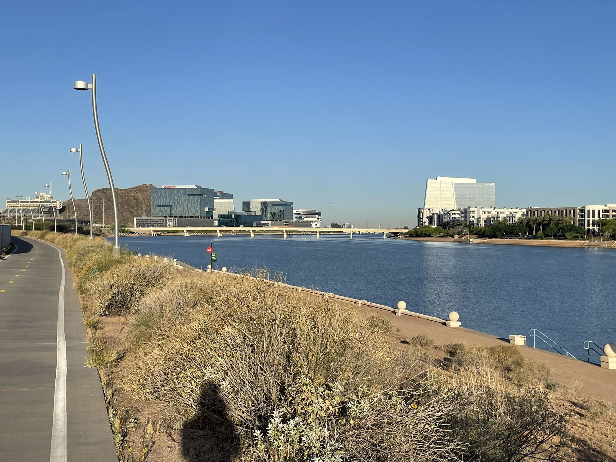 Paved path next to a lake, distant buildings, and mountains under clear sky. - Rio Salado Pathway