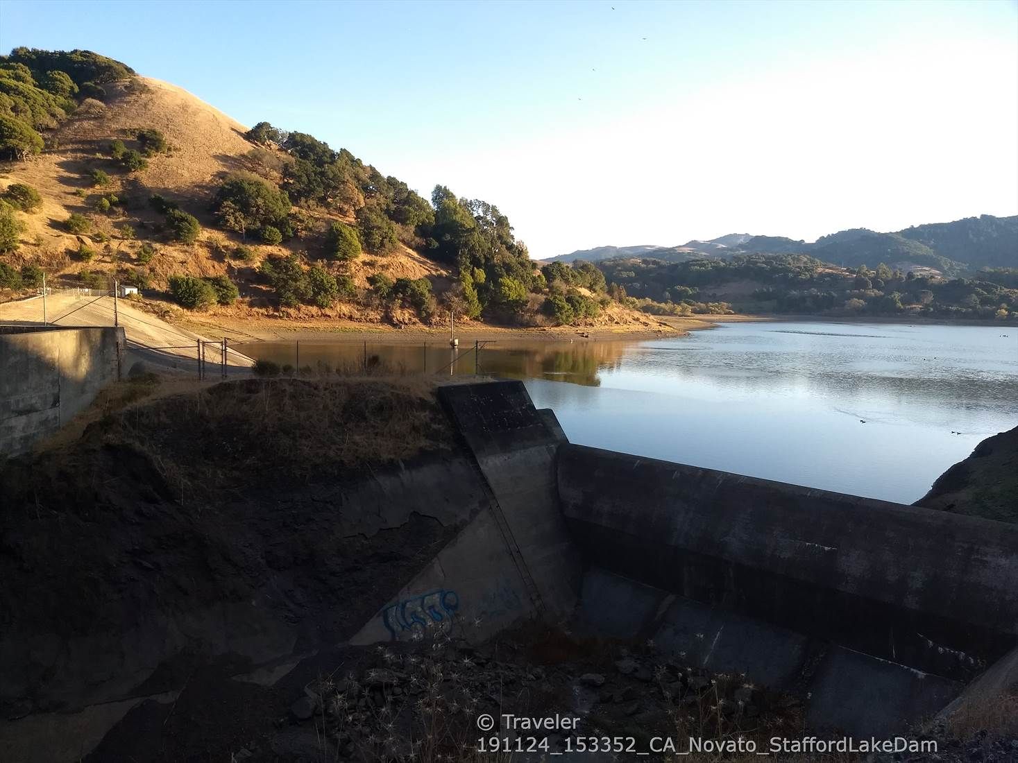 A concrete dam structure next to a lake, with dry hills under clear sky. - Enfrente Pathway – Stafford Lake Park loop from Stafford Lake