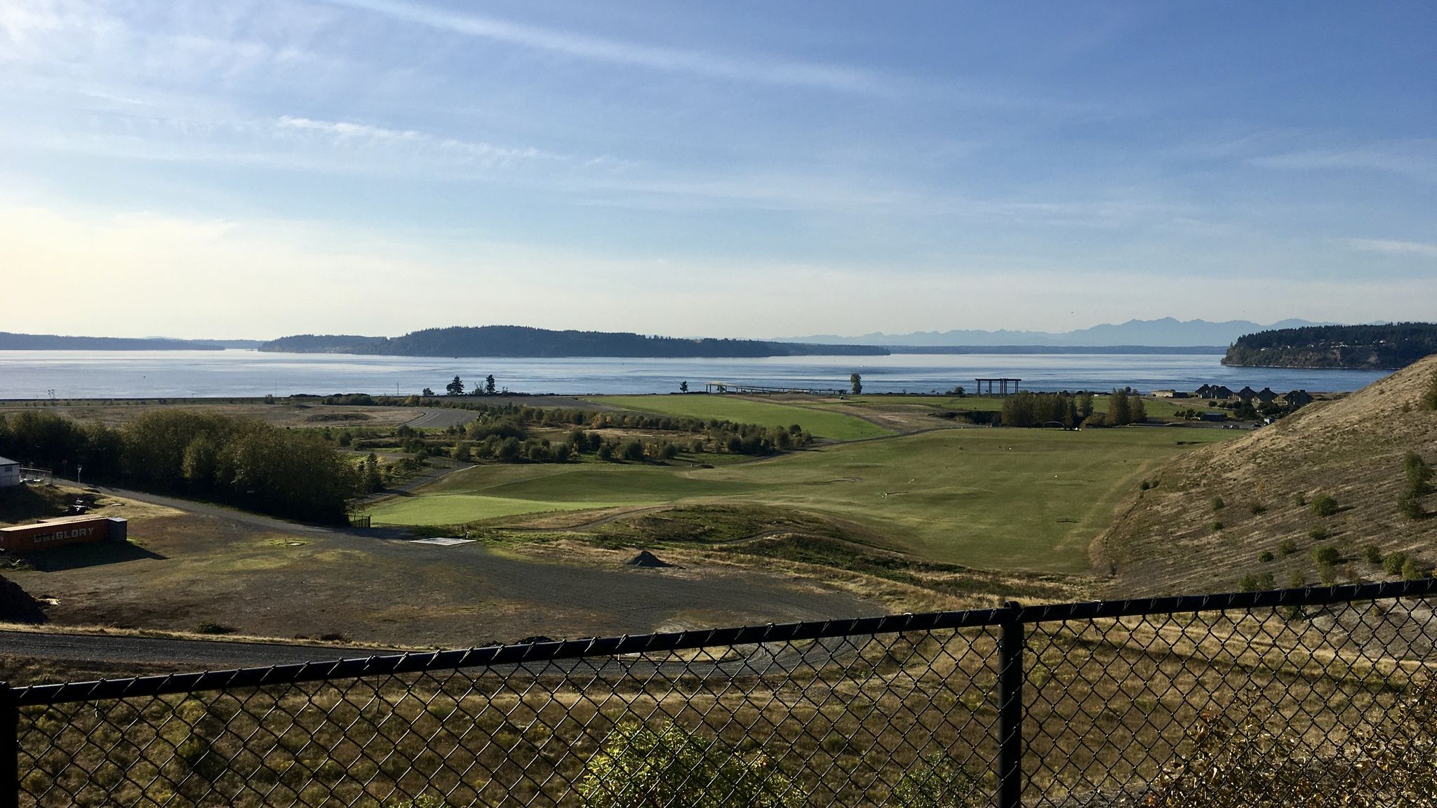 Sunny view of a green golf course, water, and hills, with a fence. - Lacey, Dupont & Lakewood loop
