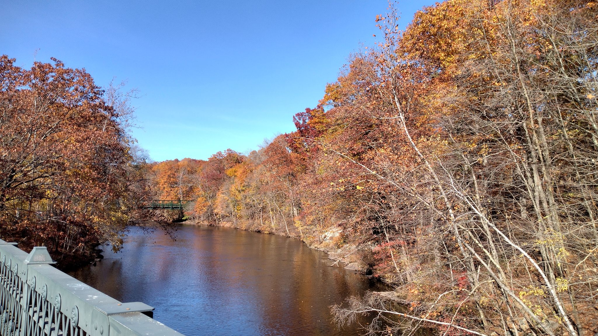 A river flows through a forest with vibrant autumn foliage under a blue sky. - Along the Blackstone River – Along the Blackstone River loop from Lincoln