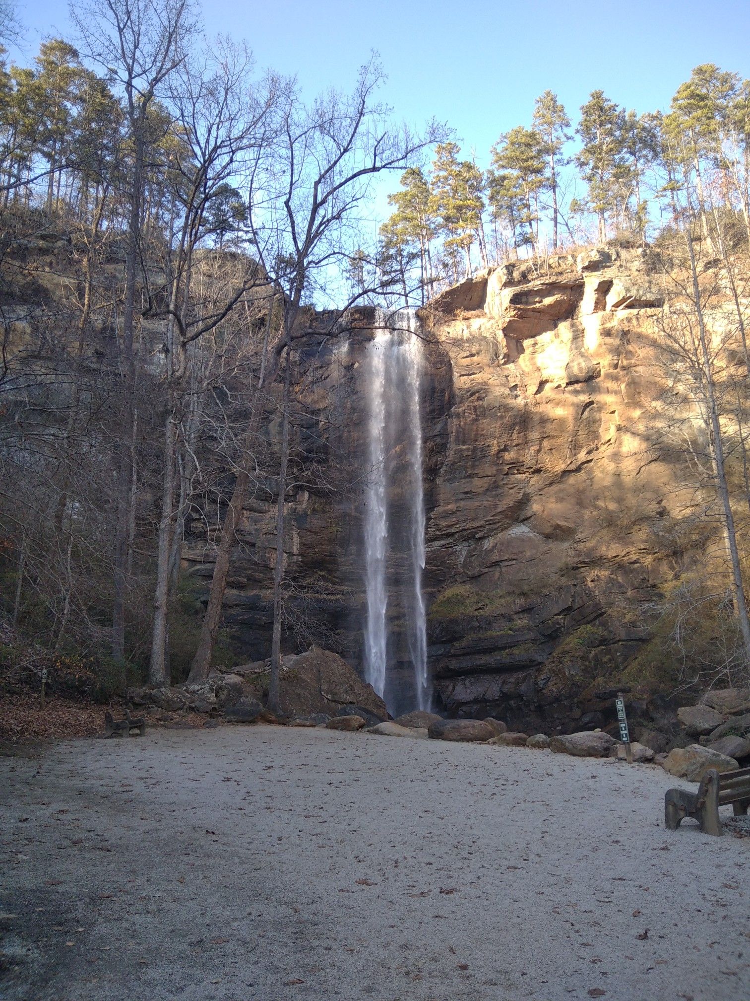 A tall waterfall flows down a cliff, with a sandy viewing area and bench. - Toccoa Falls loop from Frady Branch Trailhead Parking
