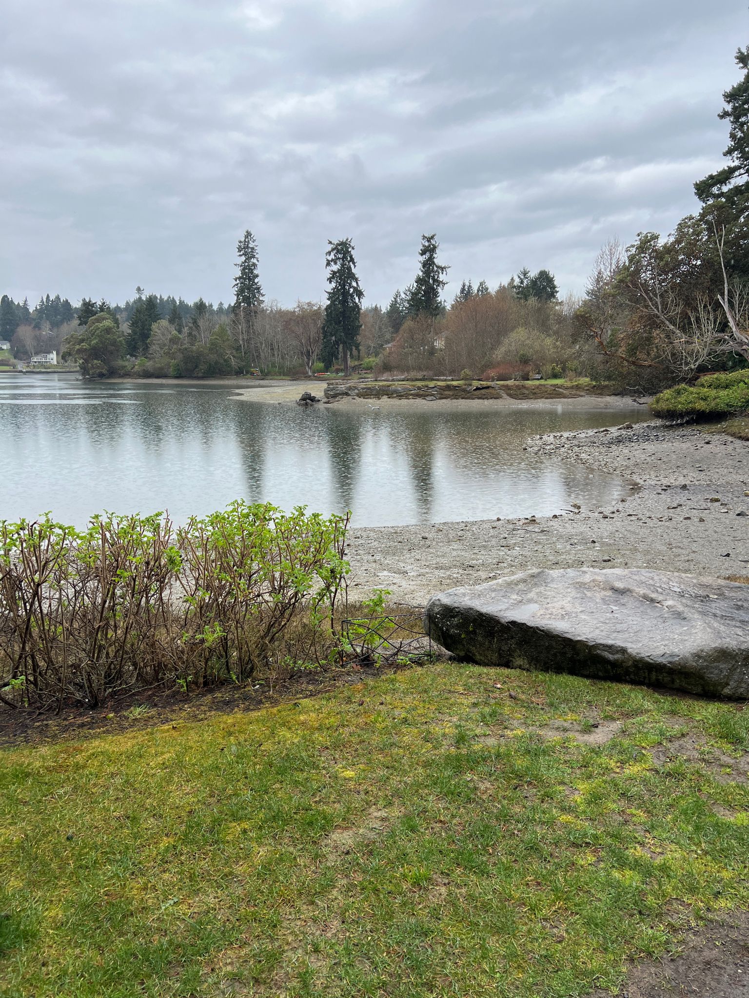 Calm water and sandy shore with a large rock under an overcast sky. - Bainbridge Waterfront Park – John Nelson Park loop from Bainbridge Island Japanese American Exclusion Memorial