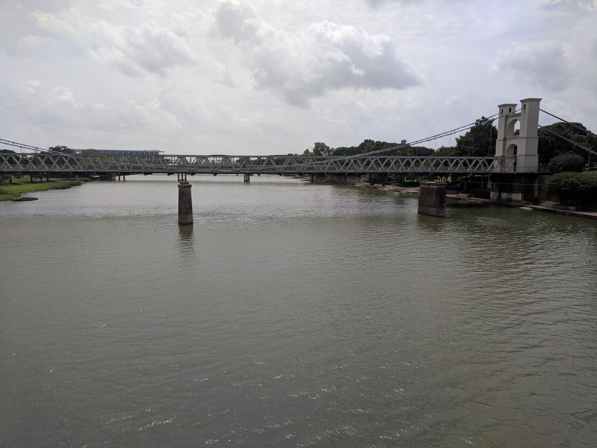 A long suspension bridge spans a wide river under an overcast sky. - Indian Spring Park Loop