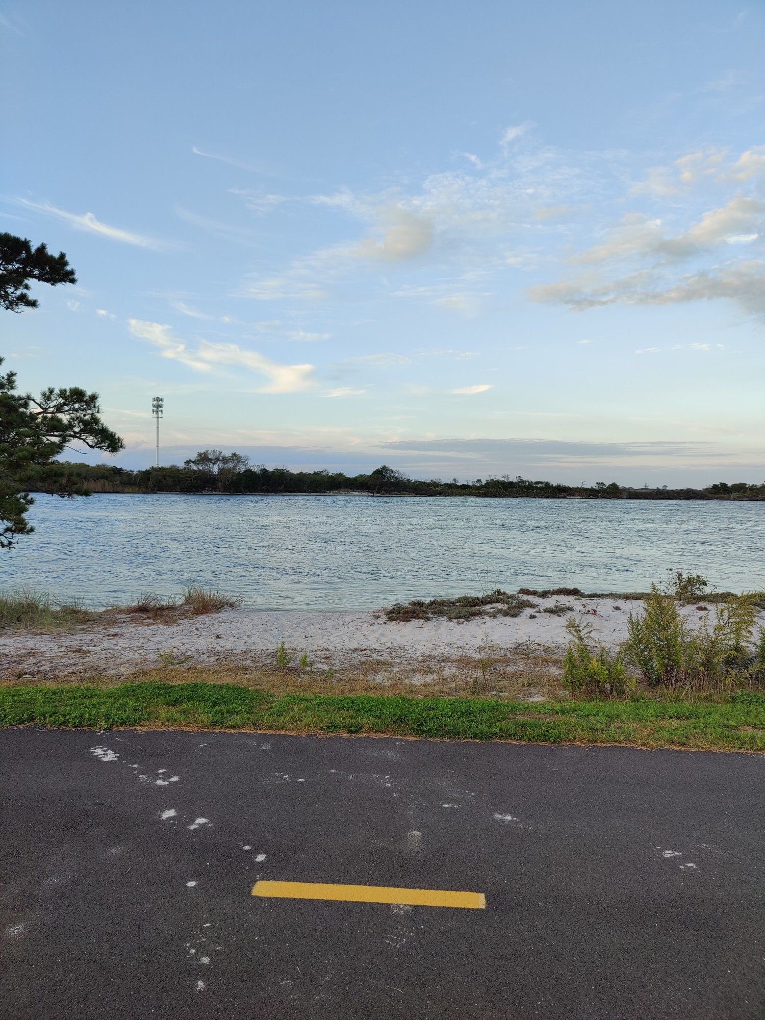 Calm water with a sandy shore, seen from a paved area under a soft sky. - Ocean Parkway Coastal Greenway loop from Captree State Park