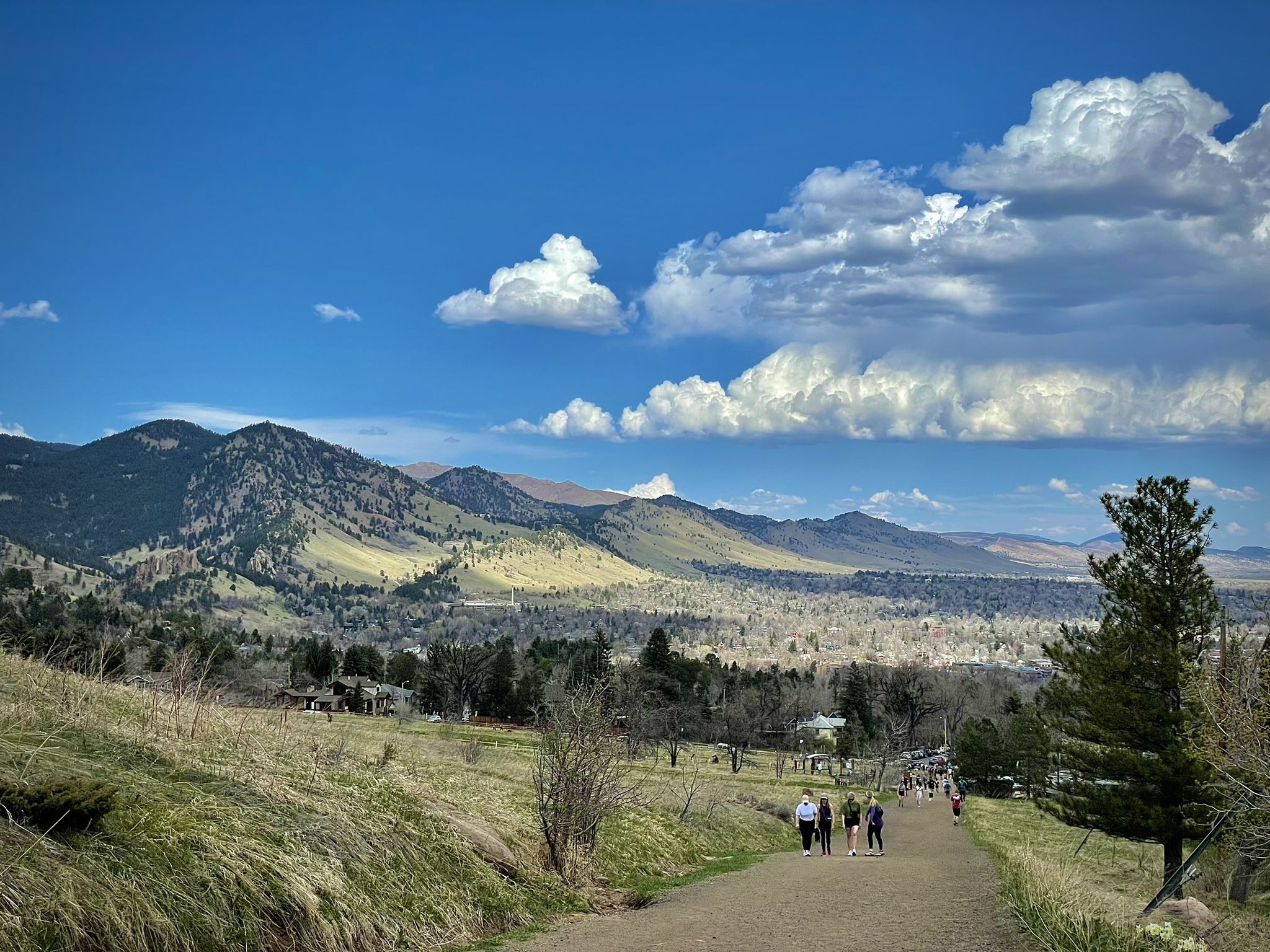 People walk on a dirt path overlooking a green valley and distant mountains under a cloudy sky. - Chautauqua and Bluebell Trail Loop