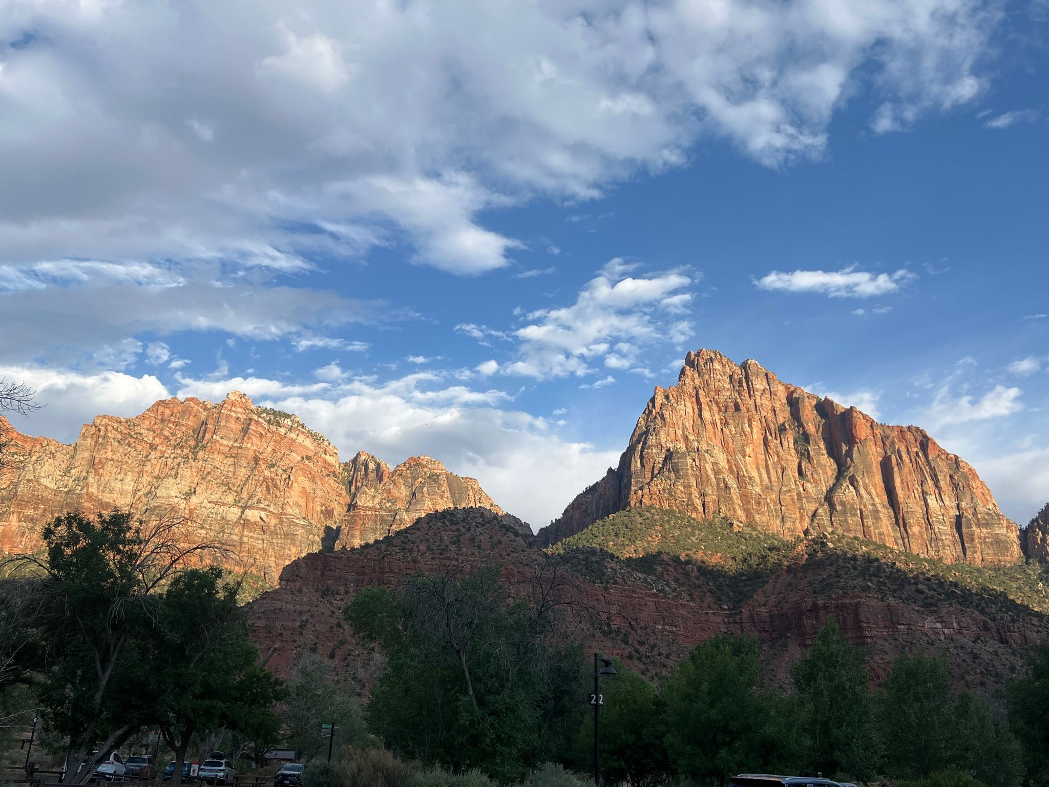 Golden hour light on towering red rock mountains, with green trees below and cloudy sky. - Watchman Trailhead – Zion National Park loop from Springdale