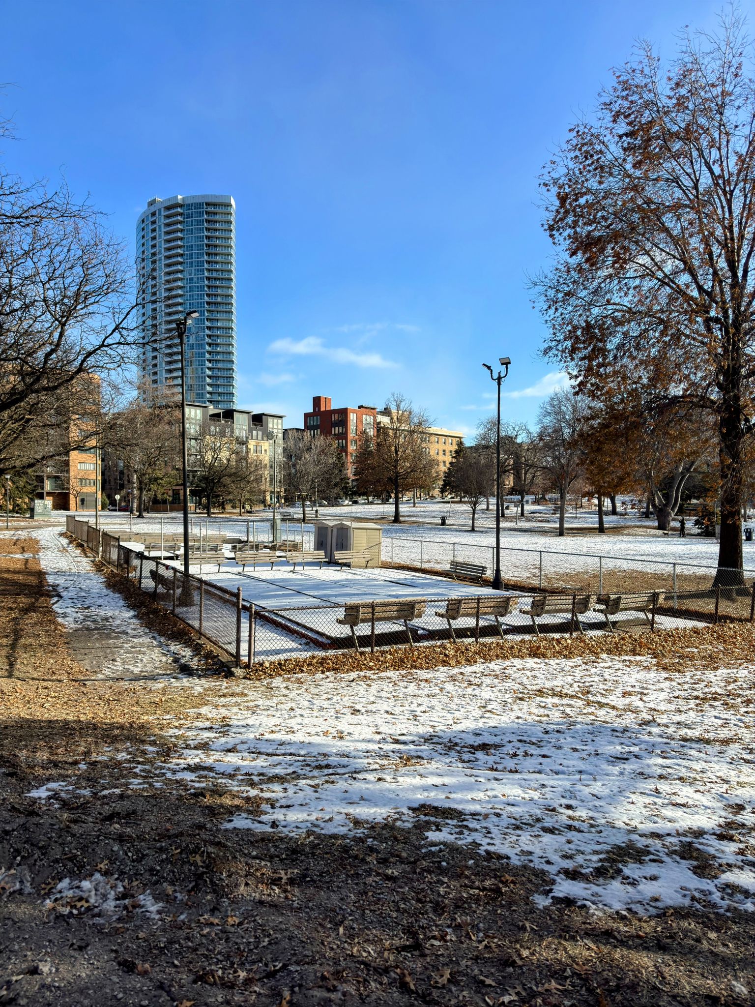 Snow-dusted park with a fenced court, benches, and city buildings in background. - Loring Park Loop