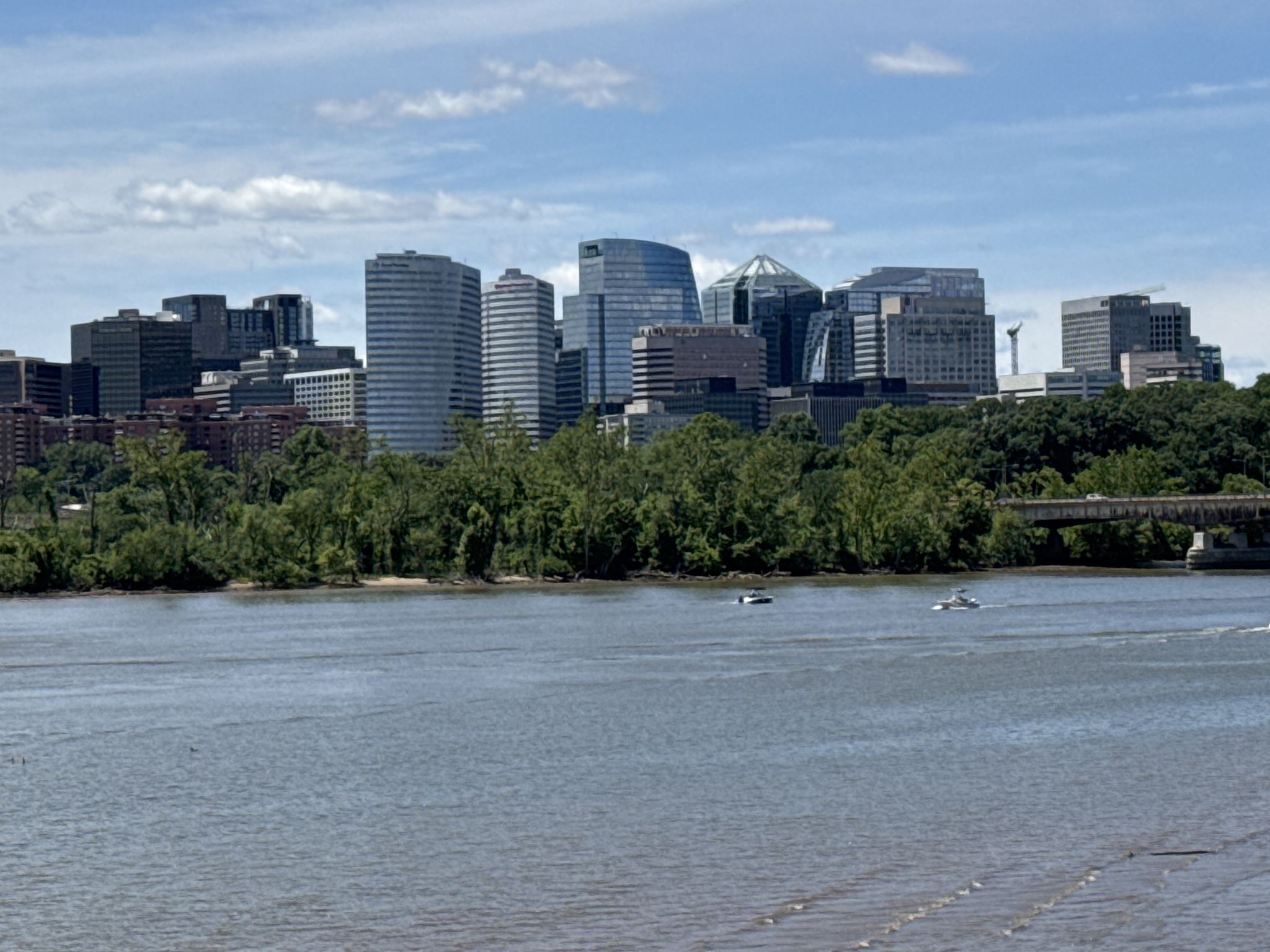A city skyline rises above a tree-lined river with small boats on a sunny day. - Netherlands Carillon – Lincoln Memorial loop from Court House